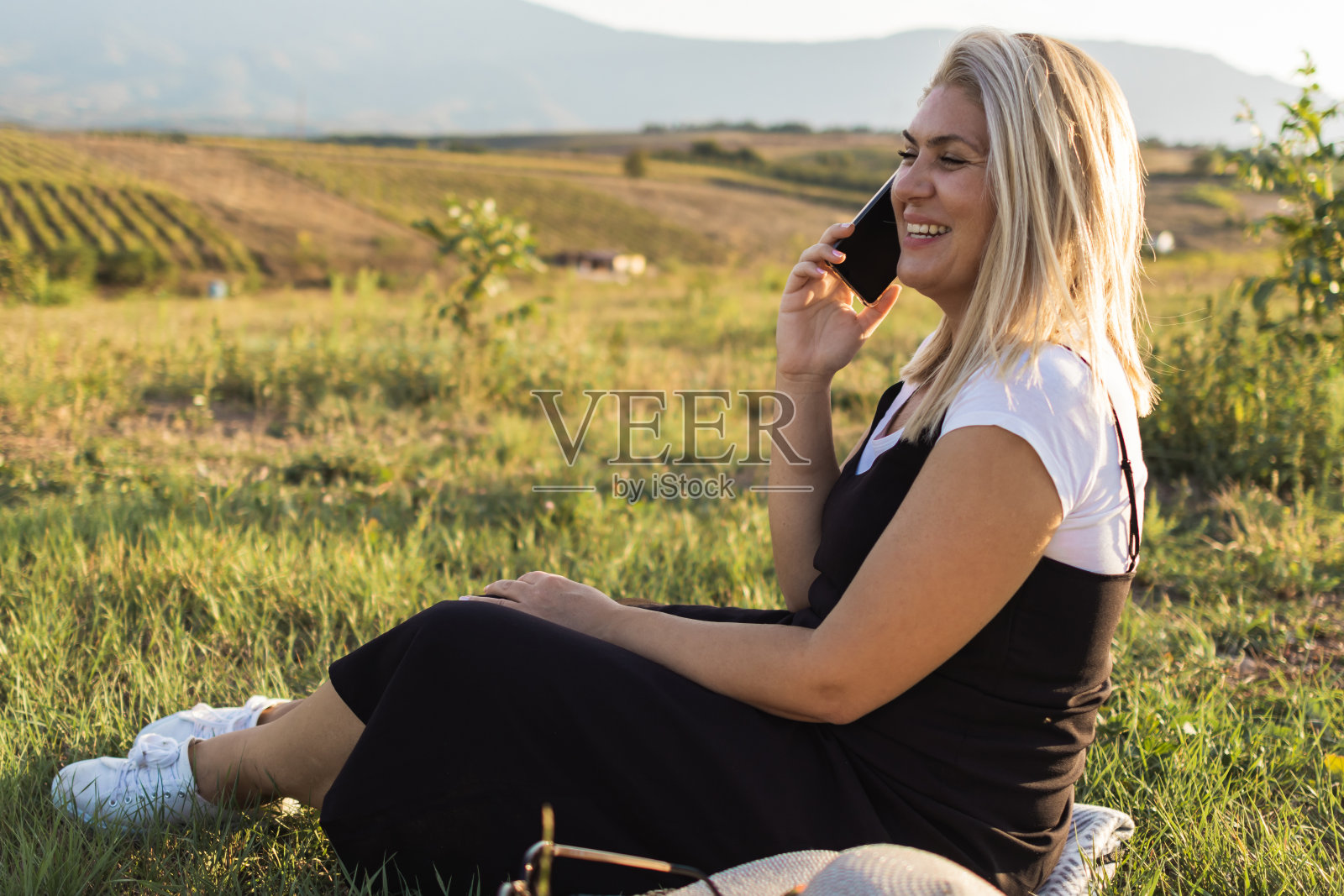 快乐的金发女人戴着帽子，享受着夏日夕阳下的草地。她正在用手机打电话。照片摄影图片