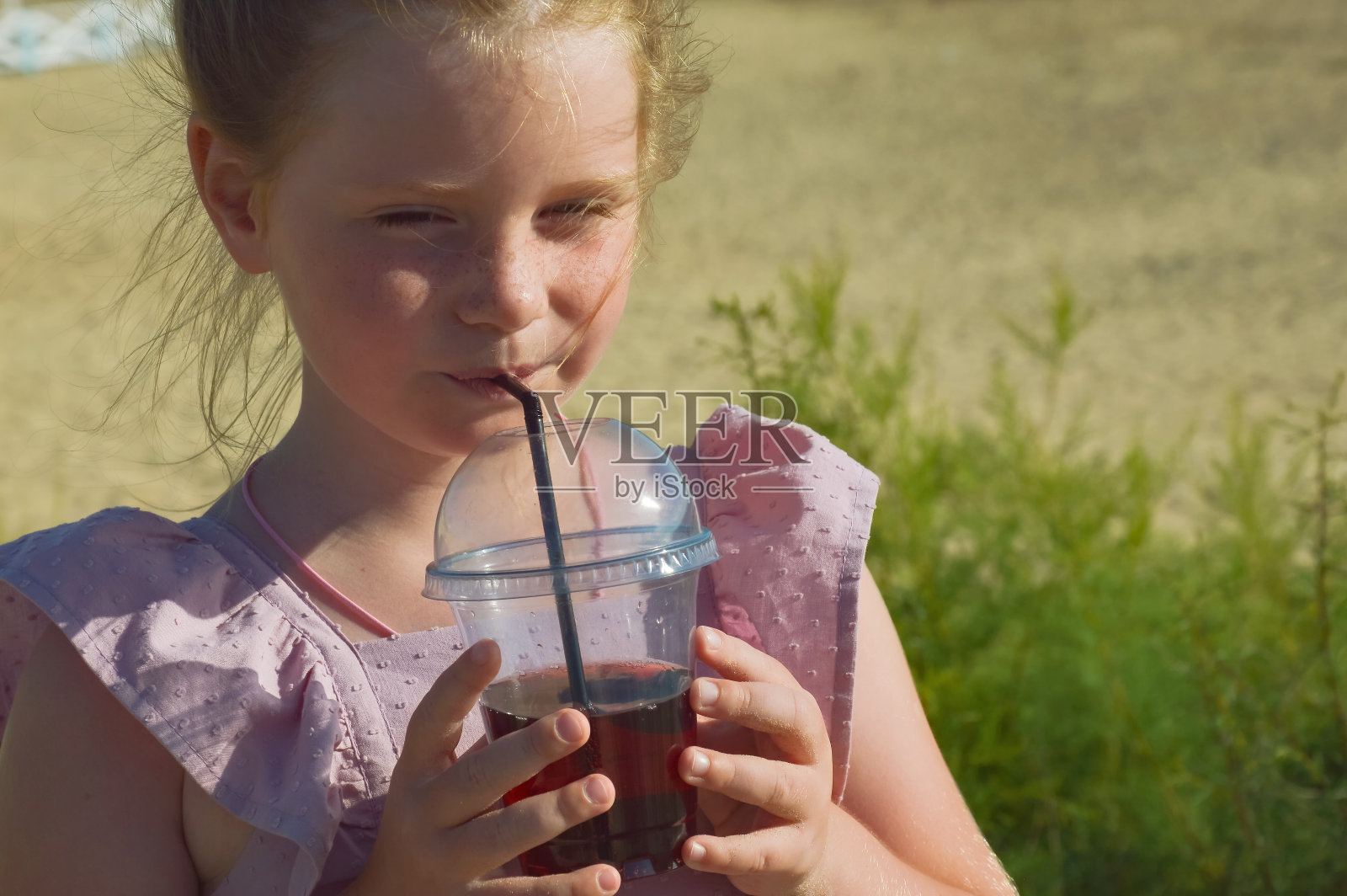 一个穿着粉红色连衣裙的小女孩在大海的背景下用吸管喝果汁。夏天阳光明媚的假期，快乐的孩子在沙滩上玩耍。设计背景。照片摄影图片