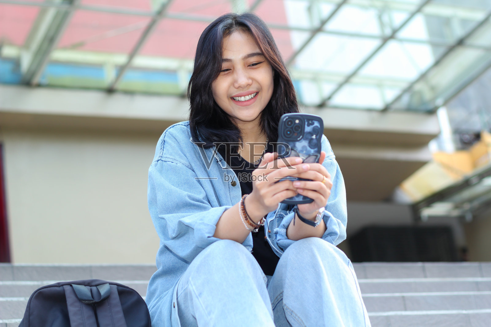 穿着休闲的亚洲年轻女性坐在户外商场的楼梯上，用智能手机应用、视频聊天、网上购物和浏览社交媒体照片摄影图片