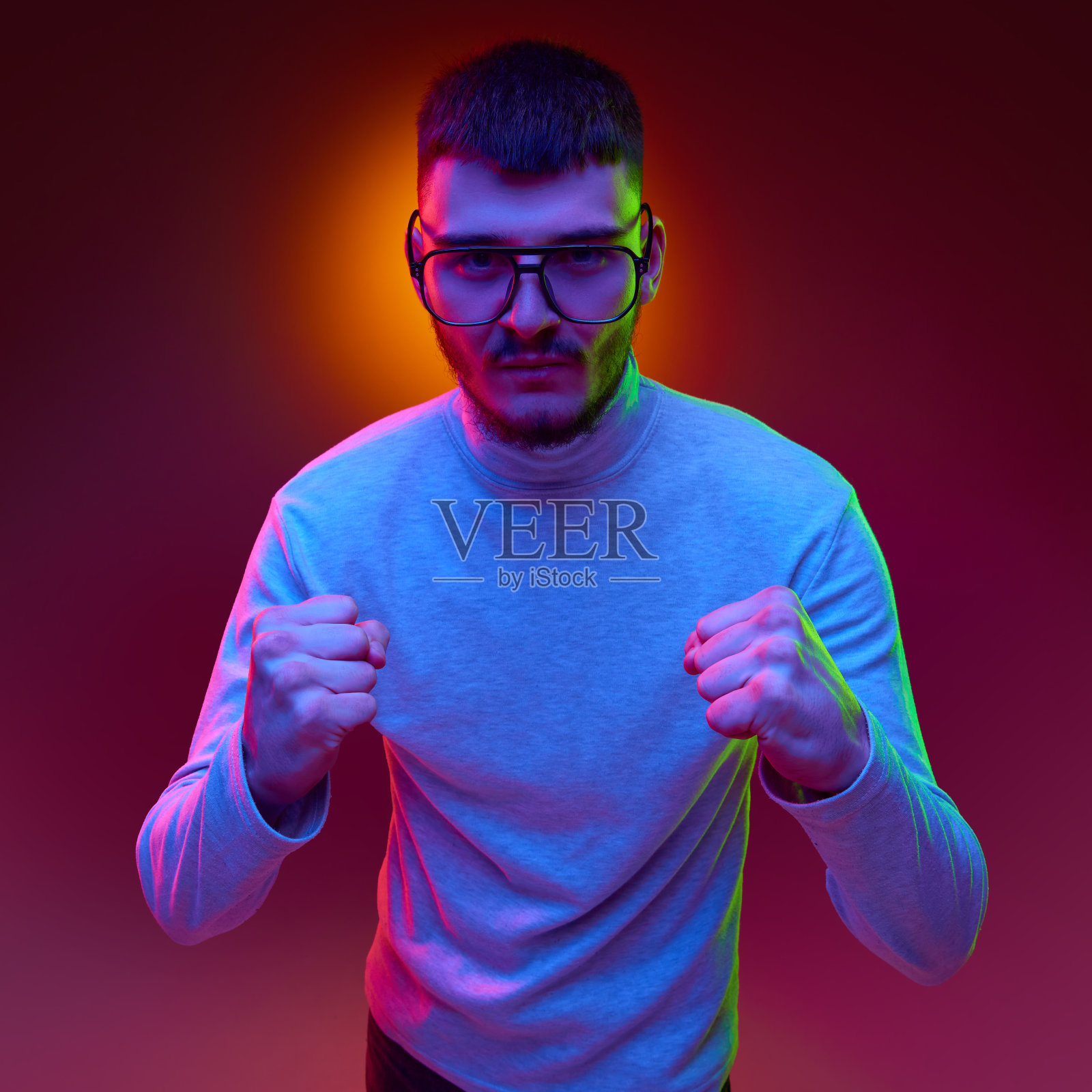 自信、严肃的年轻大胡子男子穿着随意，戴着眼镜，在混合色光和滤镜下紧握拳头看着镜头。照片摄影图片