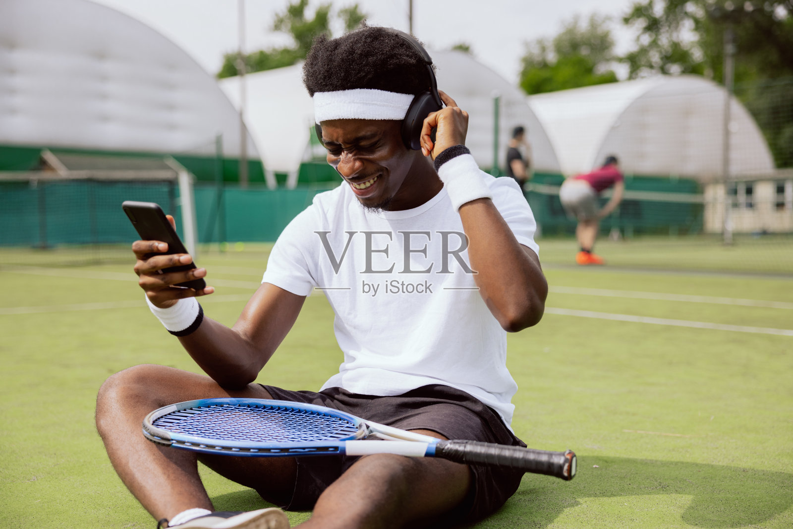 一个非洲裔的年轻人坐在网球场的草地上，戴着耳机，拿着手机，听着音乐。穿白t恤的人对这首歌很兴奋。球拍放在膝盖上照片摄影图片