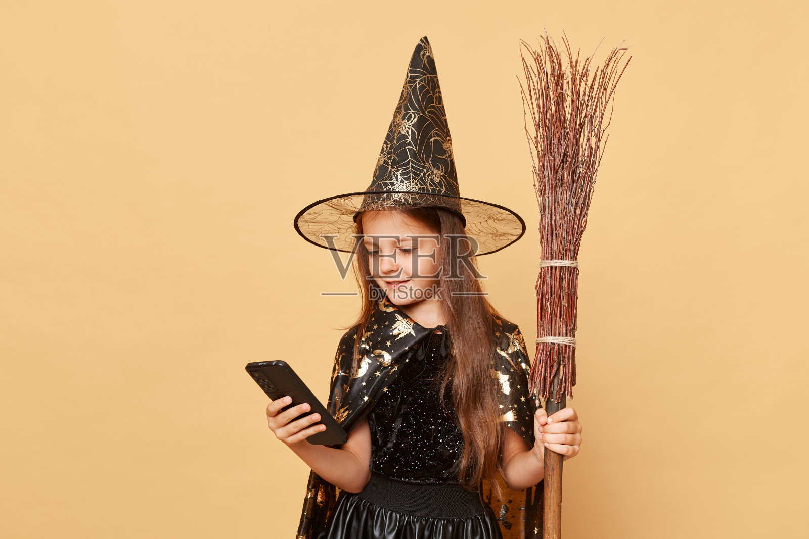 冷静可爱的小女孩穿着黑色斗篷，米色背景上有扫帚，用手机上网浏览，在线订购万圣节庆祝活动的节日道具。照片摄影图片