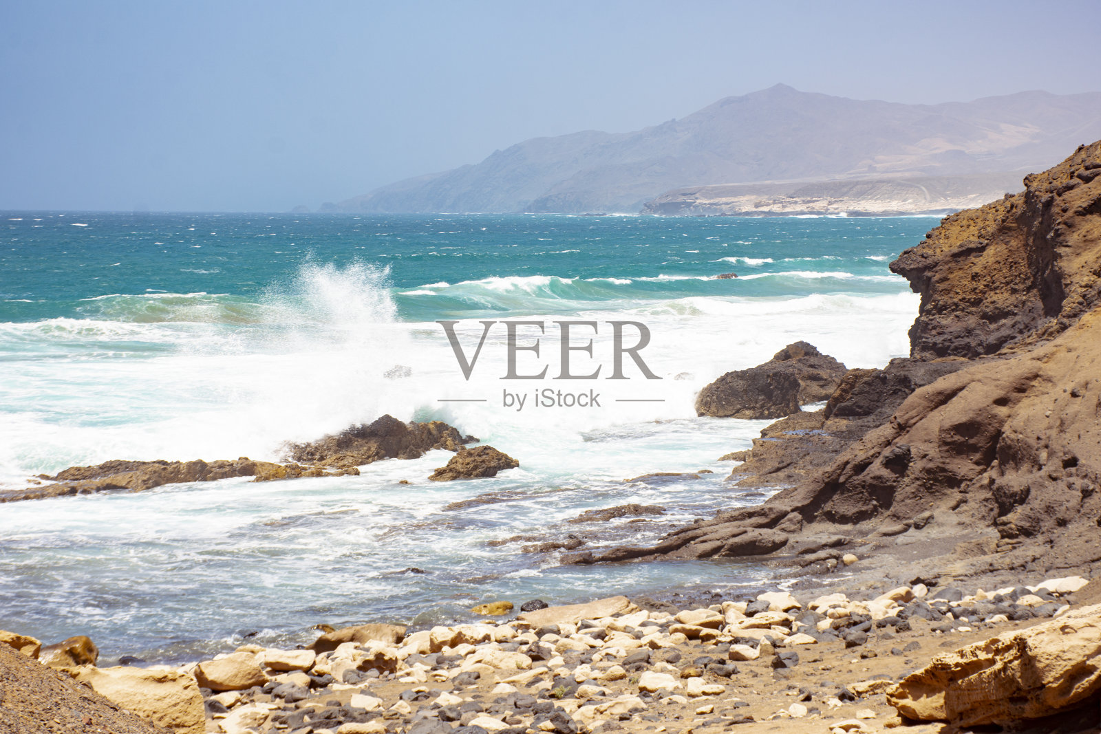 海滩的景观，强烈的海浪冲击着大海中的岩石，海岸布满了石头，背景是湛蓝天空的山脉。照片摄影图片