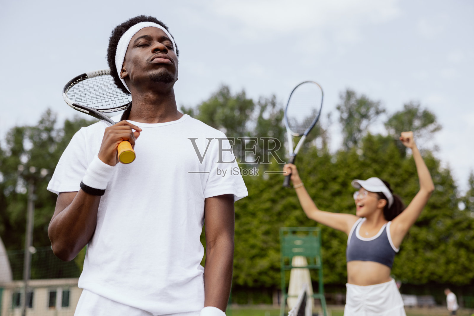 网球比赛结束后，伤心的男人输了，闭上眼睛，呼出了一口气。他把球拍挂在肩上。网球运动员穿着白色t恤，头带和腕带。在她身后，小女孩因获胜而欢欣鼓舞照片摄影图片