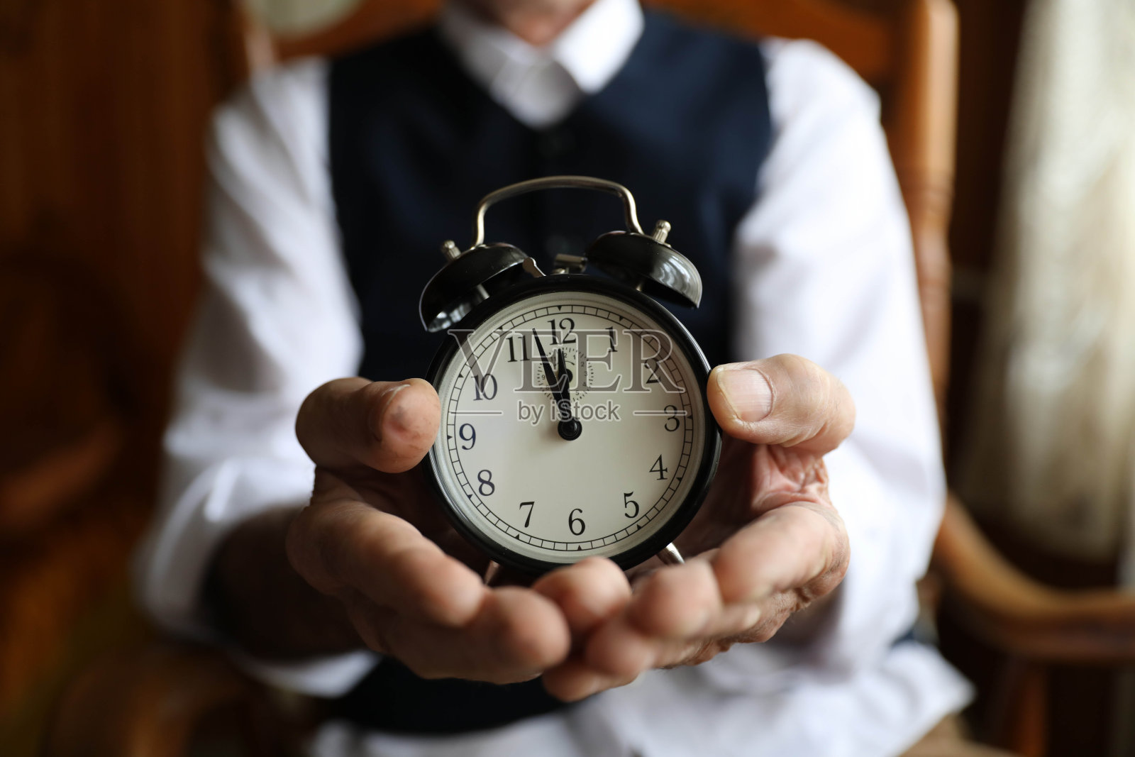 老人坐在扶手椅上，手里拿着一个小闹钟。照片摄影图片