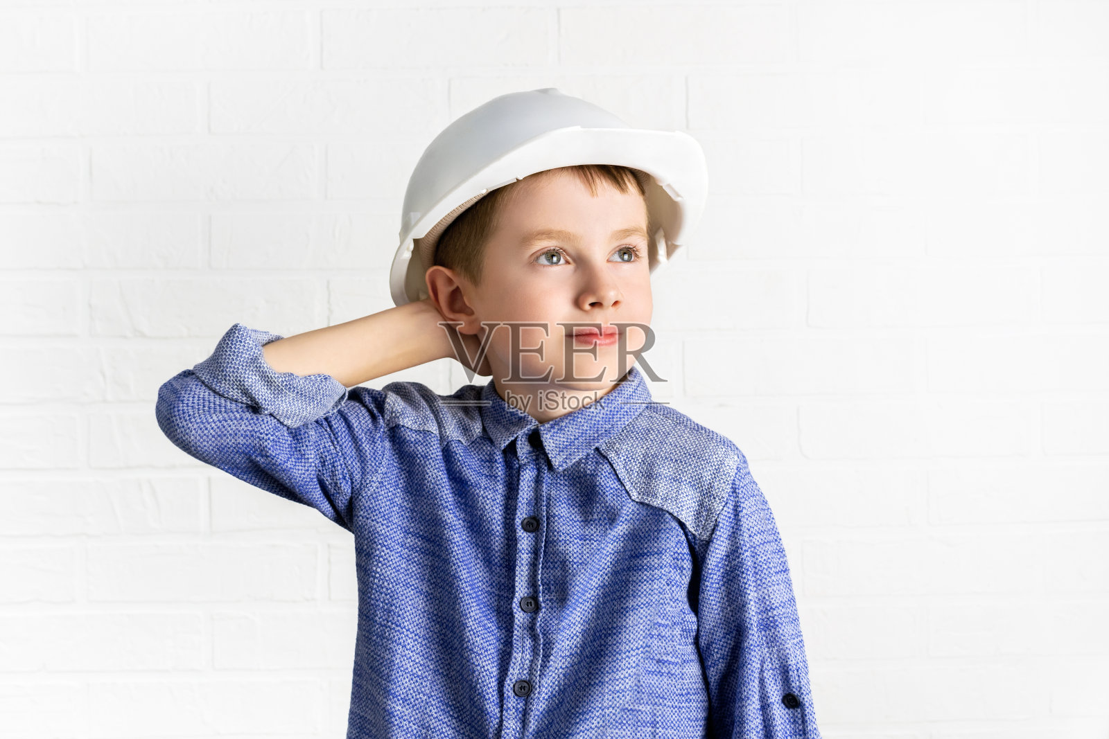 戴安全帽的孩子若有所思地抬起头，把手放在脑后。小男孩工程师，建筑师，建造者正在思考，寻找一个新的想法。照片摄影图片