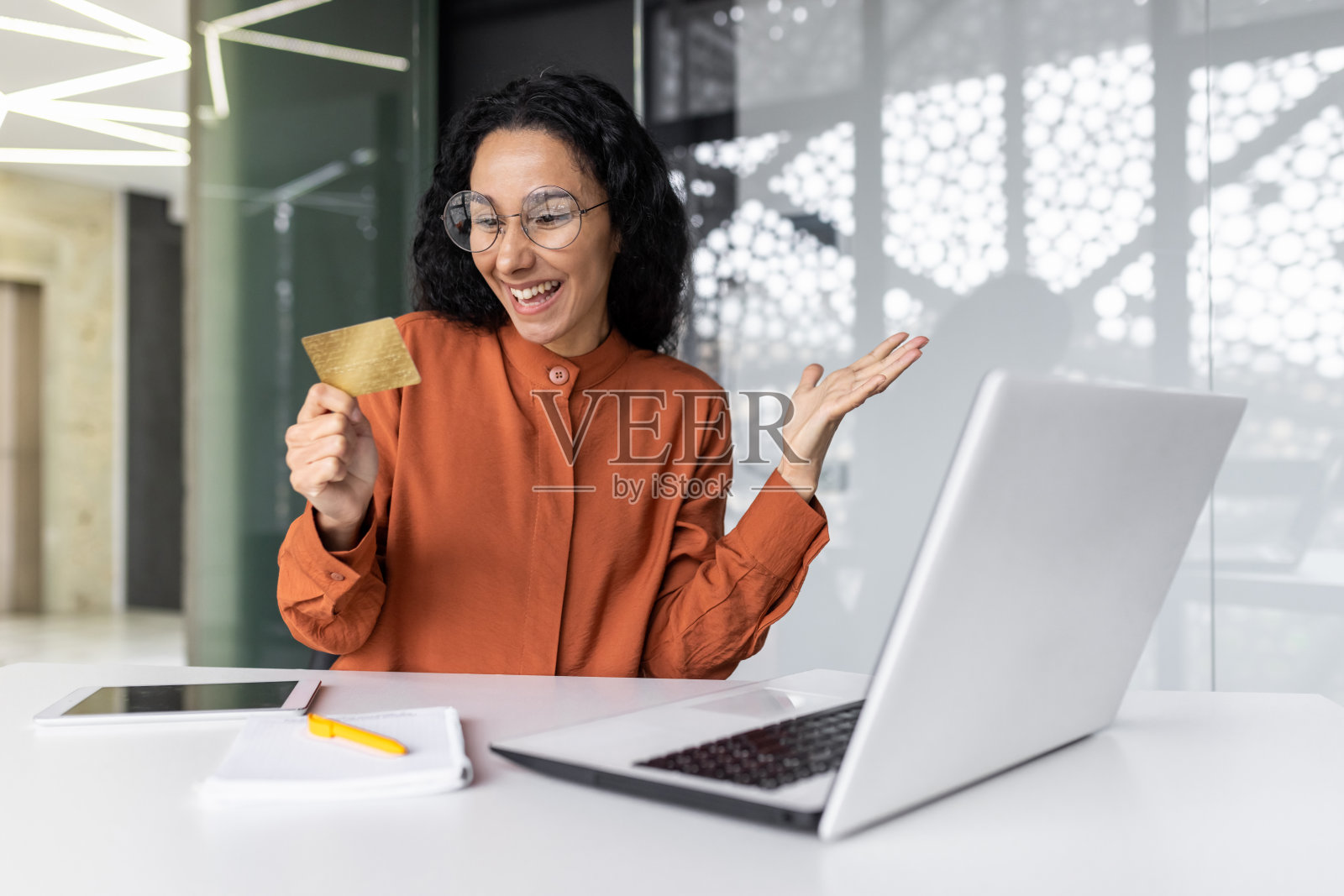 在办公室用笔记本电脑工作的年轻西班牙裔女子，手持信用卡微笑照片摄影图片