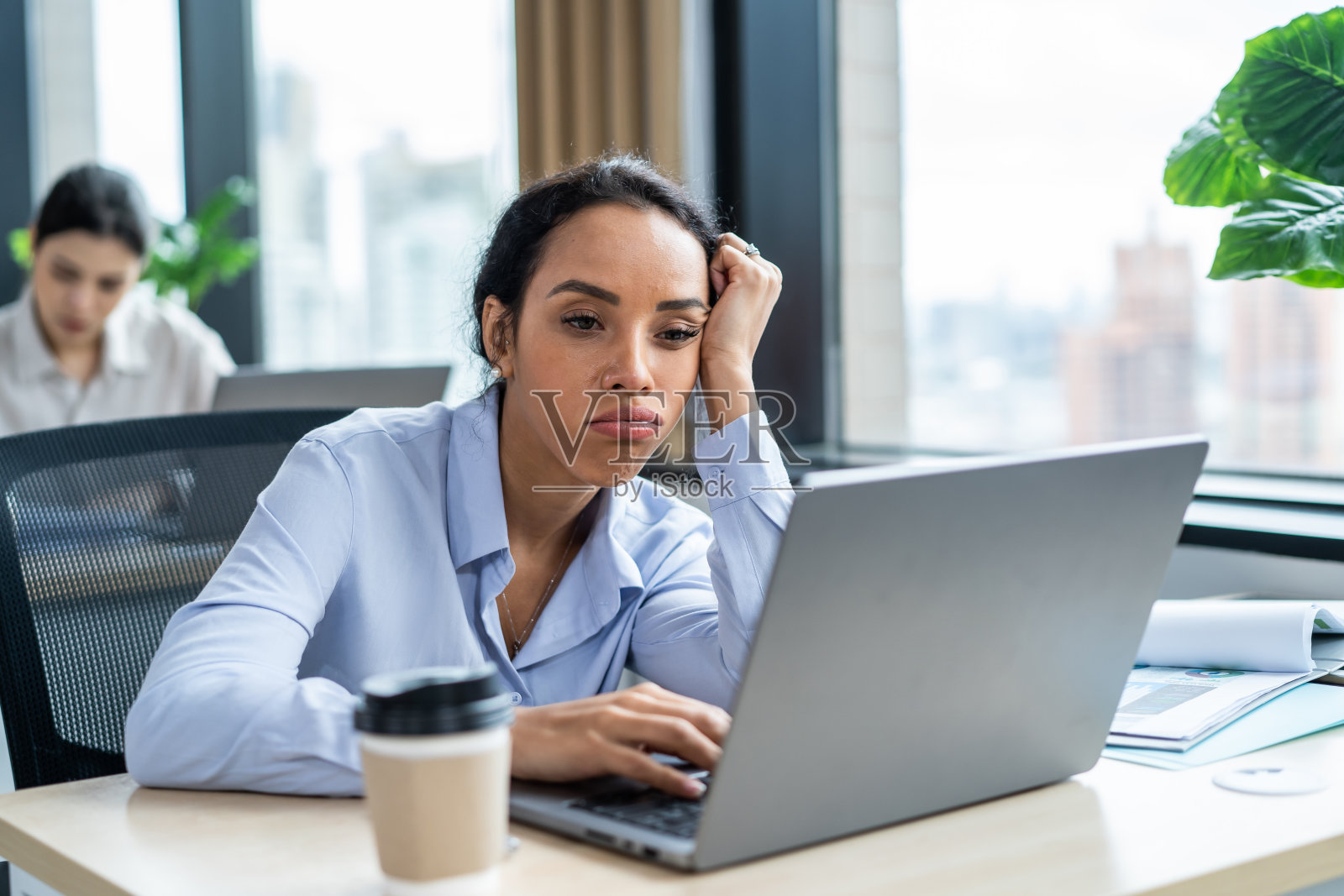 筋疲力尽的拉丁裔年轻女商人在办公室工作过度。迷人的忙碌的女员工感到疲倦和困倦，然后在桌子上喝咖啡，用电脑在公司计划项目照片摄影图片