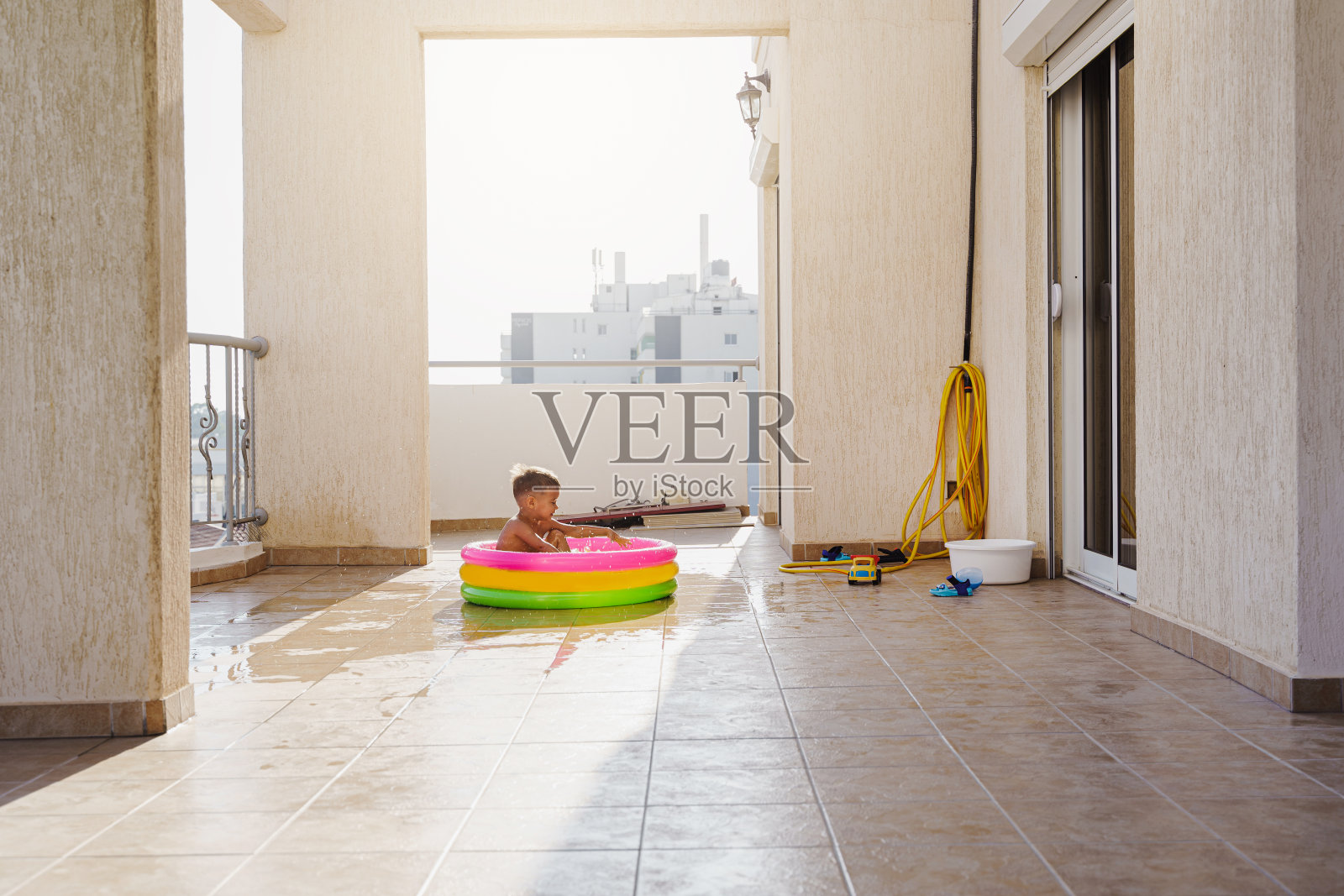 在炎热的夏日里，一个蹒跚学步的男孩在阳台上的充气圆形游泳池里欢快地玩耍照片摄影图片