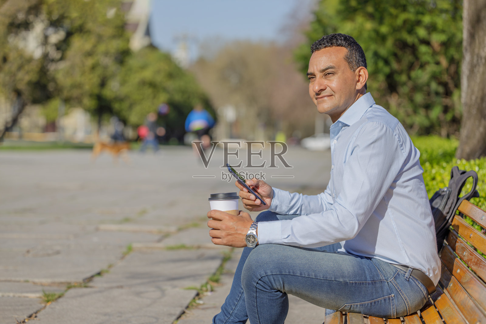 穿着衬衫的拉丁裔男子的肖像，拿着一杯一次性咖啡坐在公园的长凳上。照片摄影图片