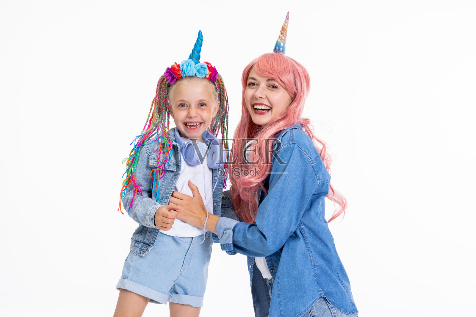 欢快的年轻母亲和学龄前的女儿戴着独角兽发带，戴着粉红色的假发，穿着牛仔衣服，站在白色的背景上，在工作室摆姿势照片摄影图片
