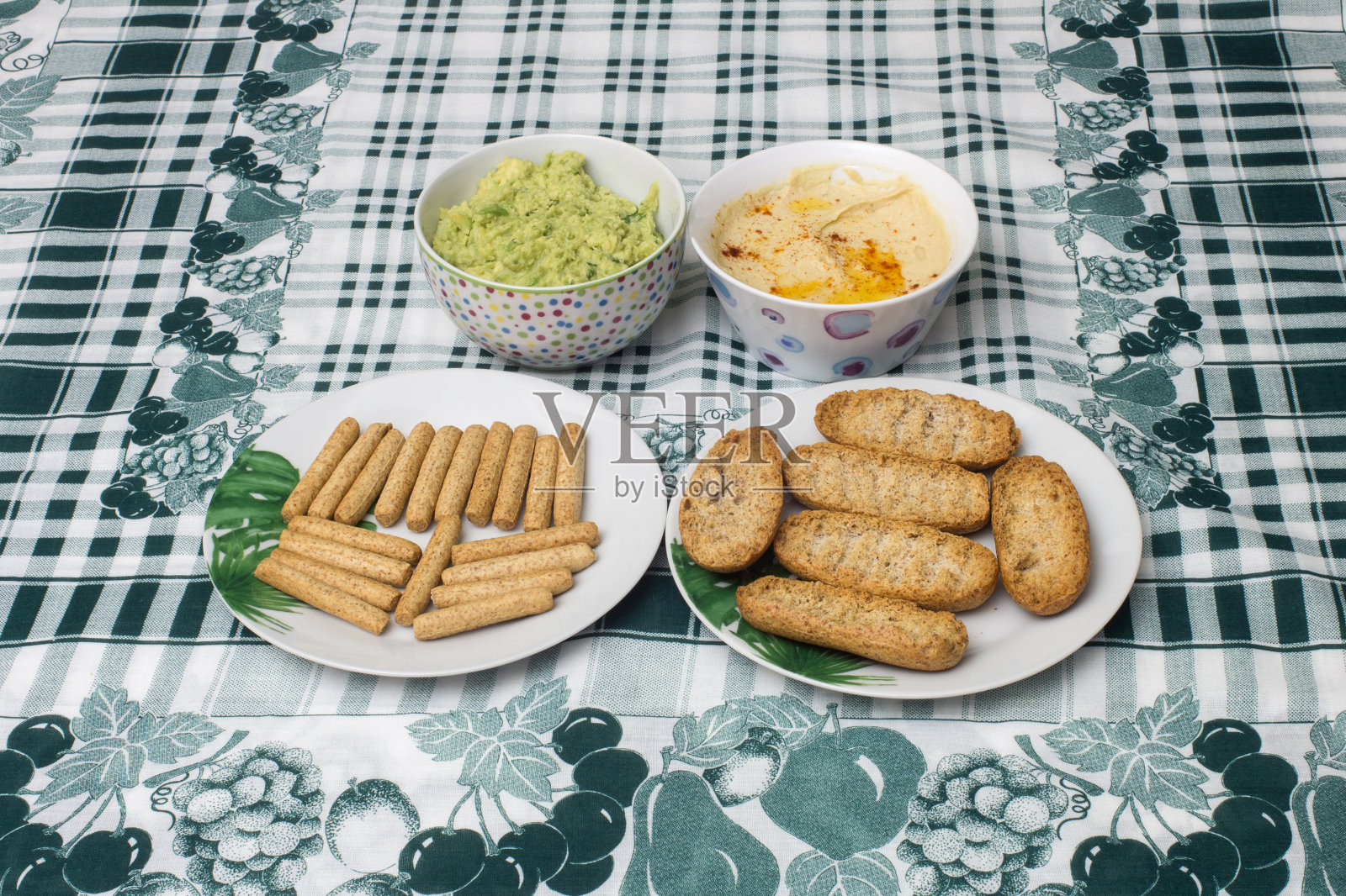 两小碗自制的鹰嘴豆泥和鳄梨酱，配有配有全麦吐司和面包条的盘子，上面铺着时尚的布桌布照片摄影图片