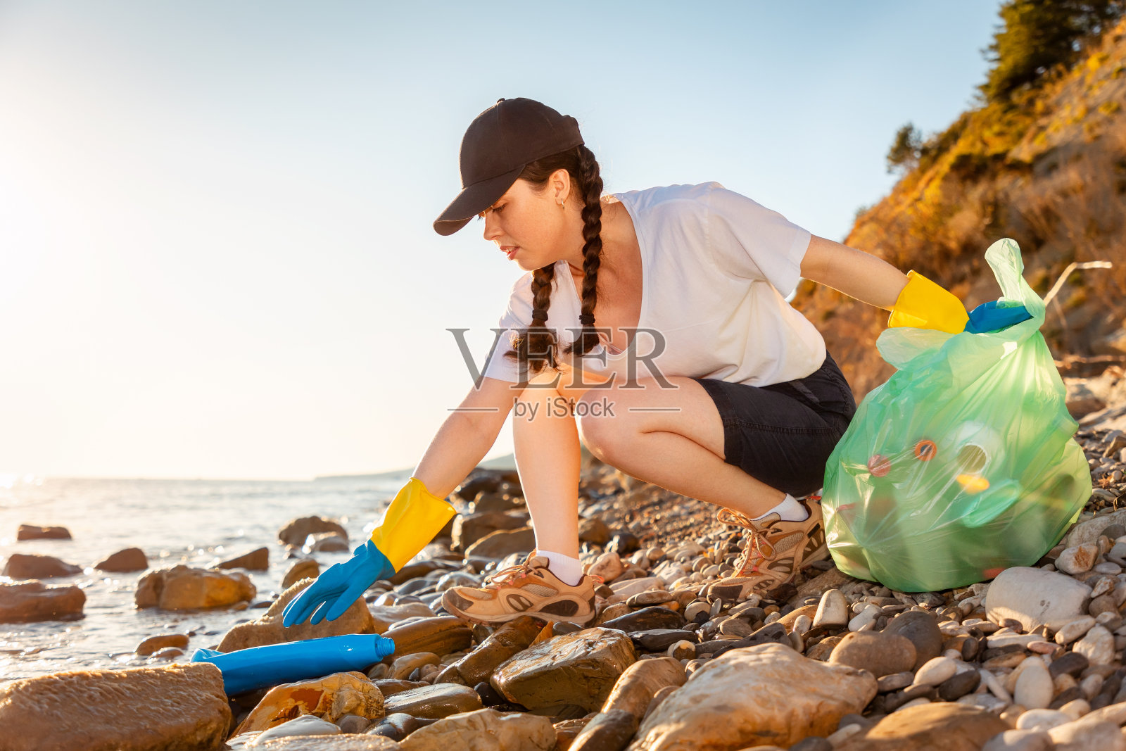 戴着橡胶手套的年轻白人女子蹲在卵石沙滩上收集垃圾的侧视图。地球日和环境灾难的概念照片摄影图片