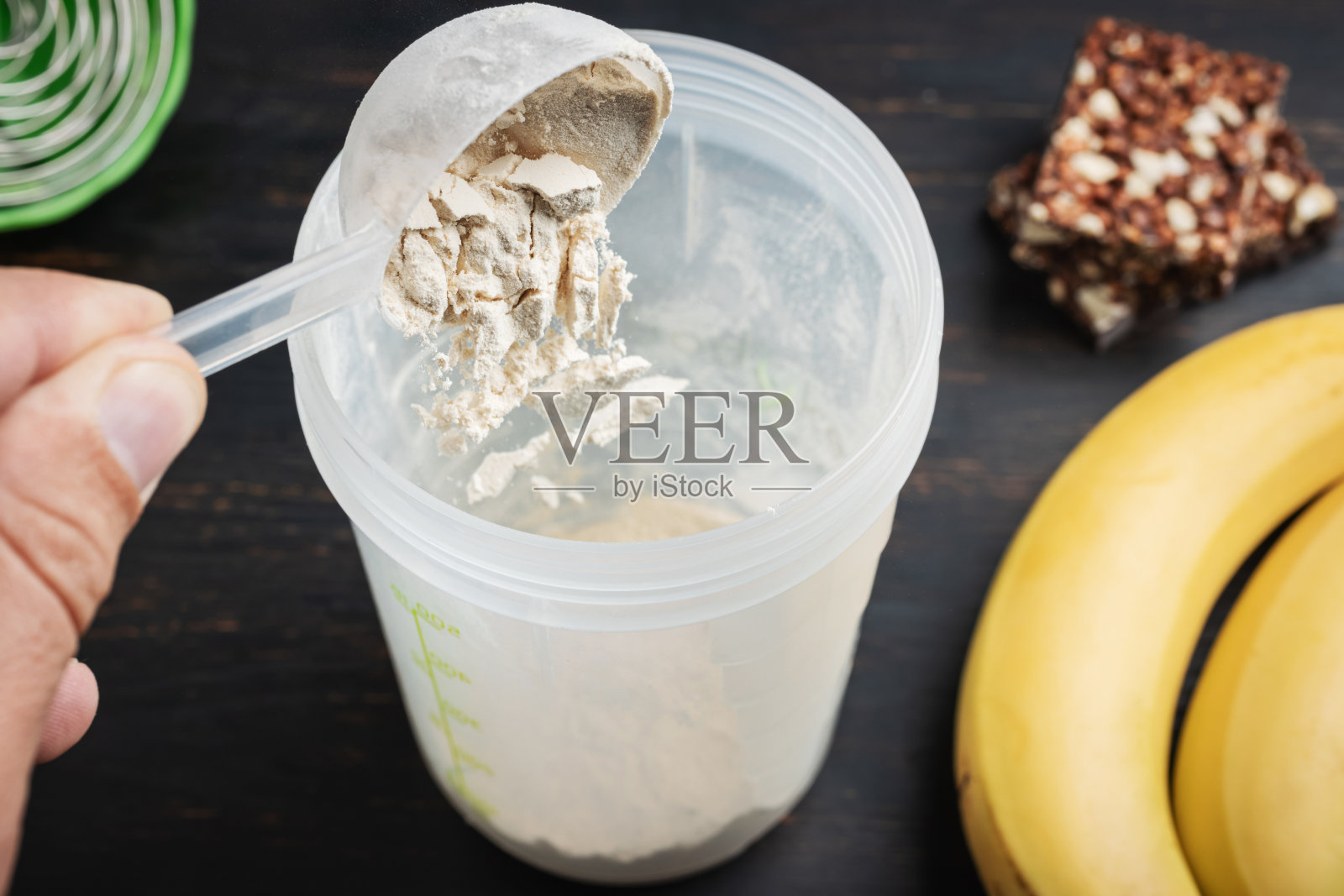 男手放入摇壶舀取部分乳清、大豆蛋白粉。蛋白质巧克力棒和香蕉放在一块深色木板上，是健身食品的补充照片摄影图片