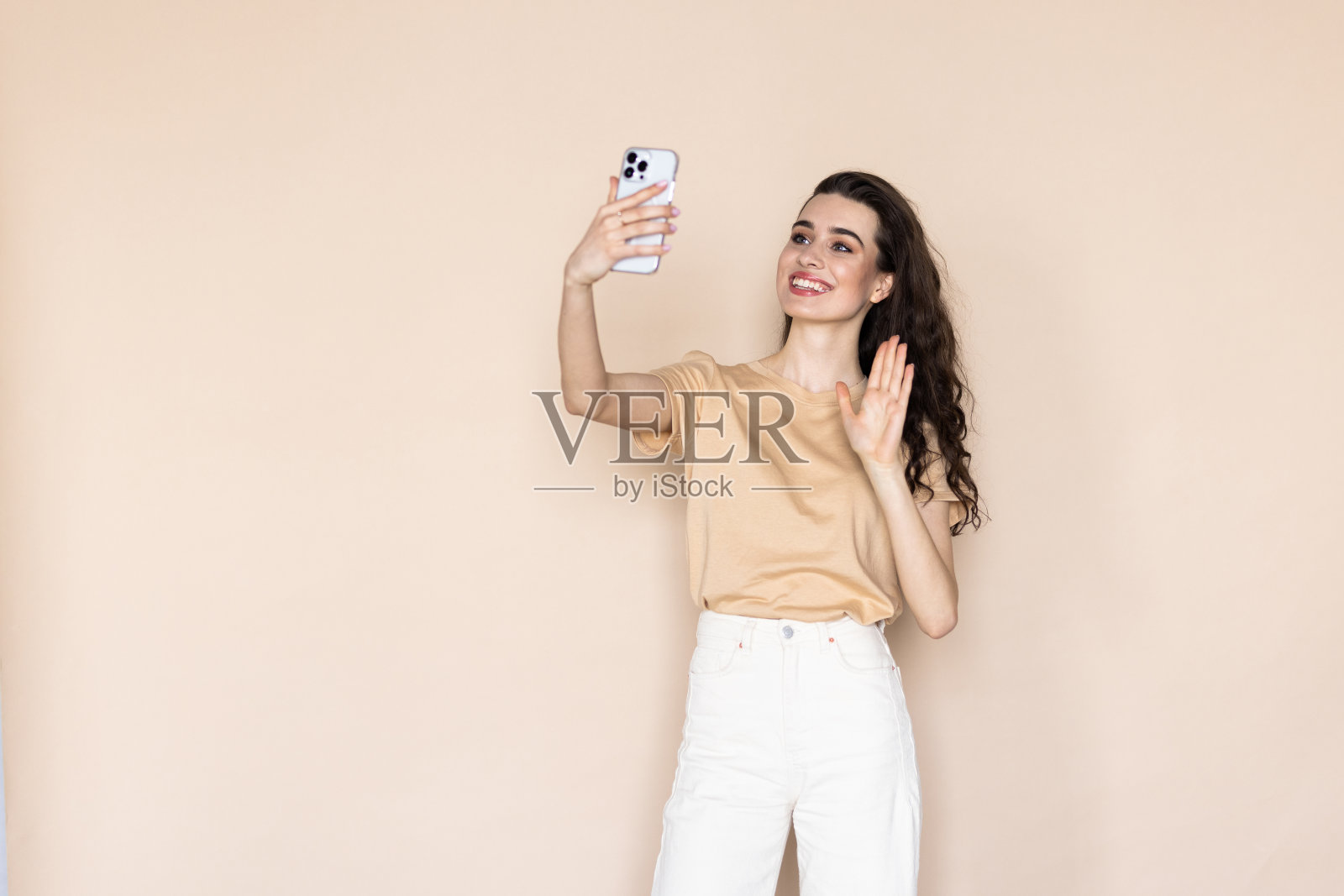 米色背景的年轻女子在电话上挥舞着她的手，正在通过视频通话照片摄影图片
