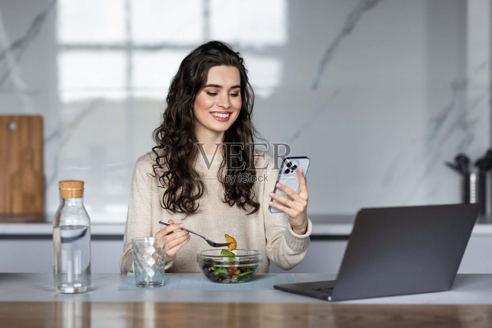 漂亮的运动女人一边吃着一碗沙拉，一边在家里的厨房用手机发短信。照片摄影图片