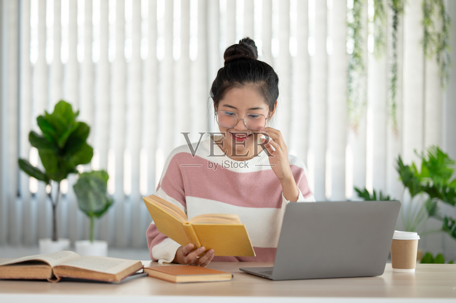 一位戴着眼镜的快乐漂亮的亚洲女大学生正在书桌前看书。照片摄影图片