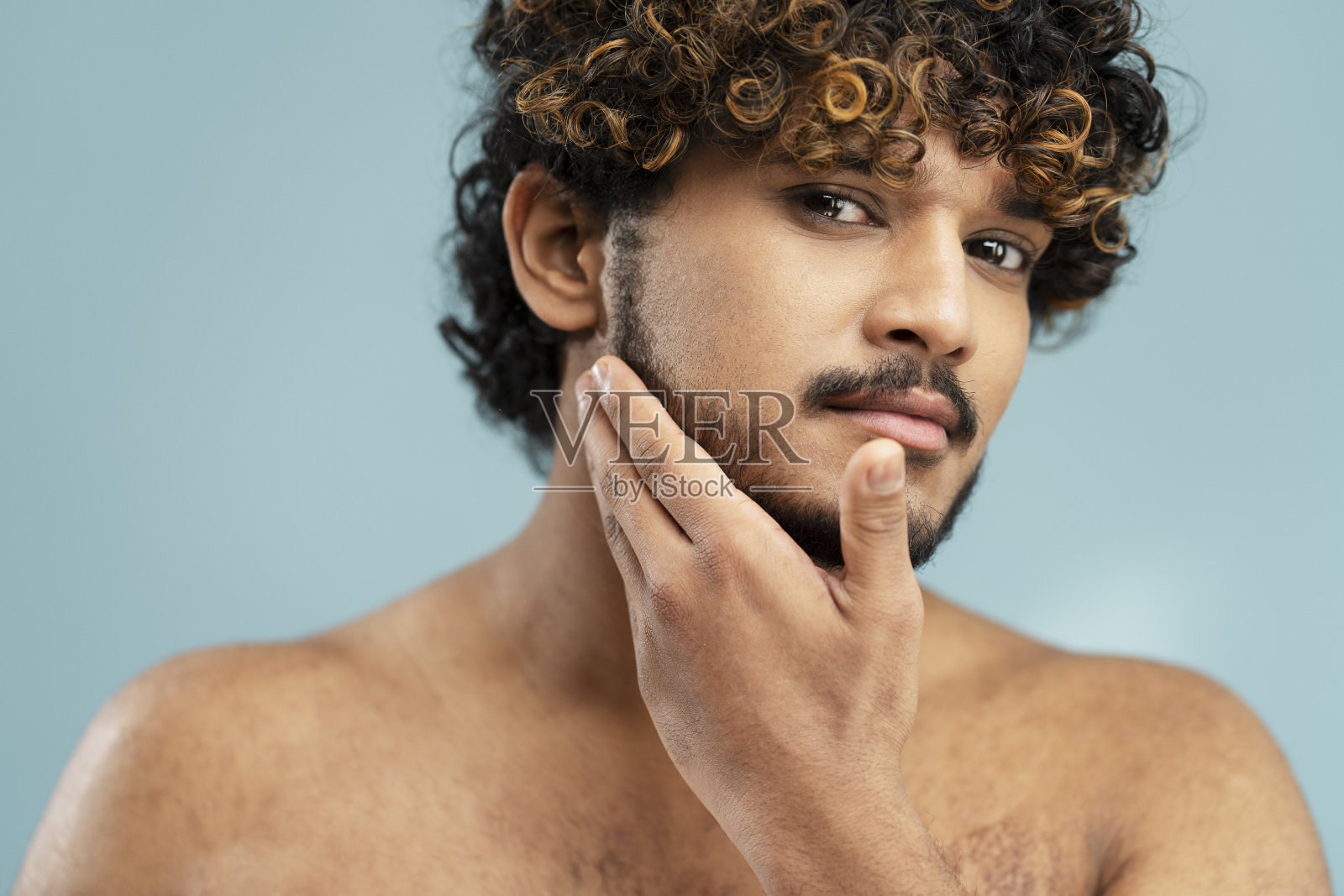在蓝色背景下，年轻的印度男子享受着清新光滑的皮肤，触摸着胡须照片摄影图片