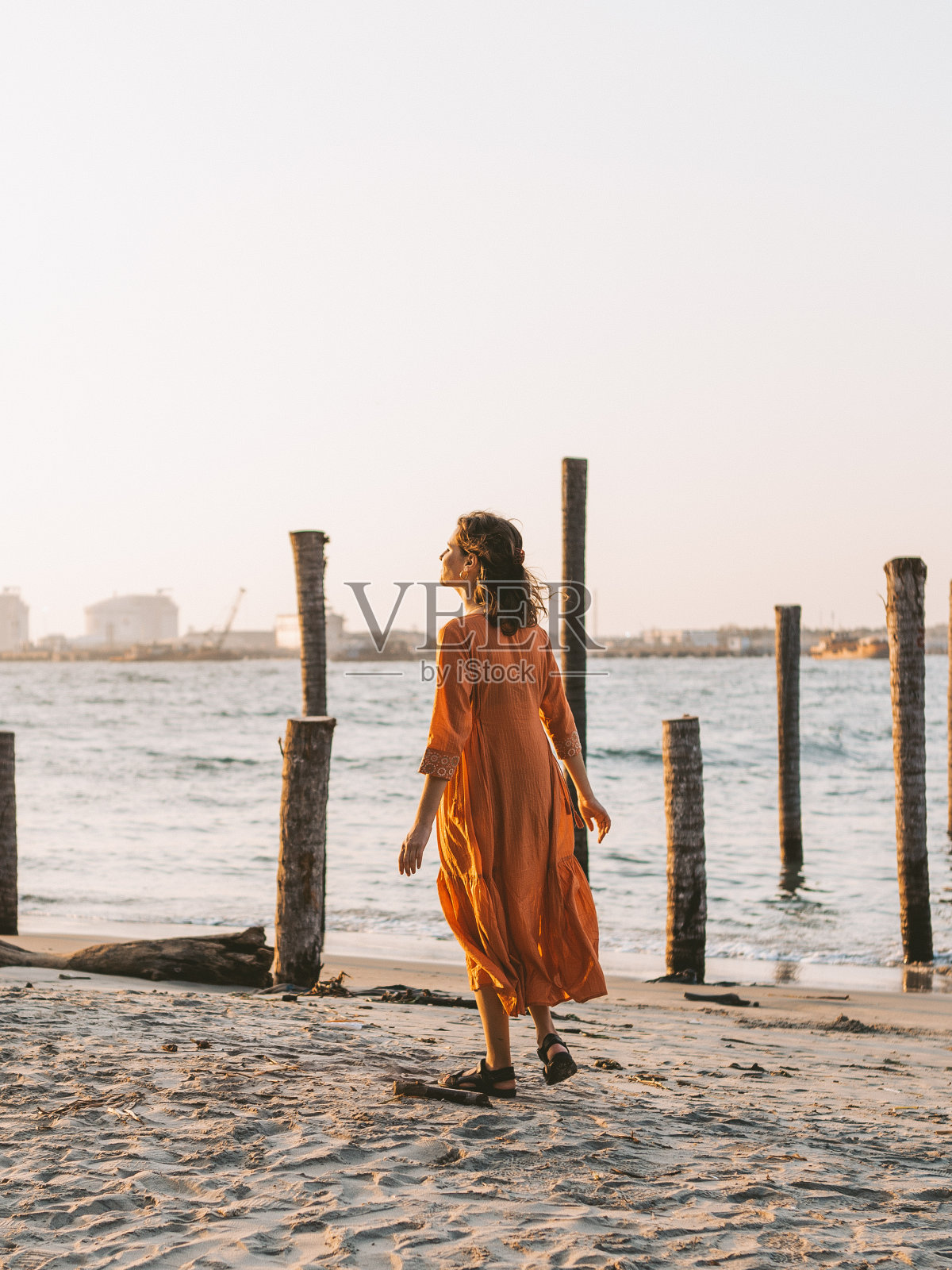 一位身穿橙色连衣裙的年轻女子站在海滩上。一些木头矗立在水面上，这是在日落的时候。照片摄影图片