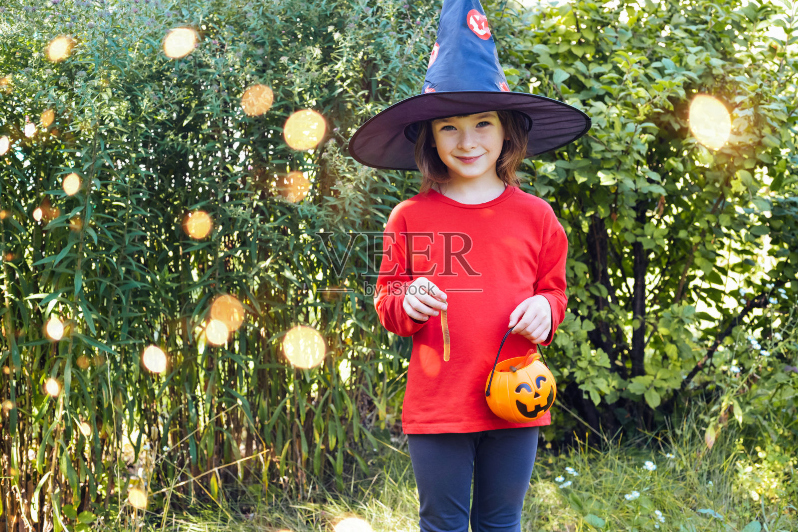 戴着狂欢节女巫帽的女孩手里拿着一个南瓜篮子，里面装着糖果和软糖。照片摄影图片