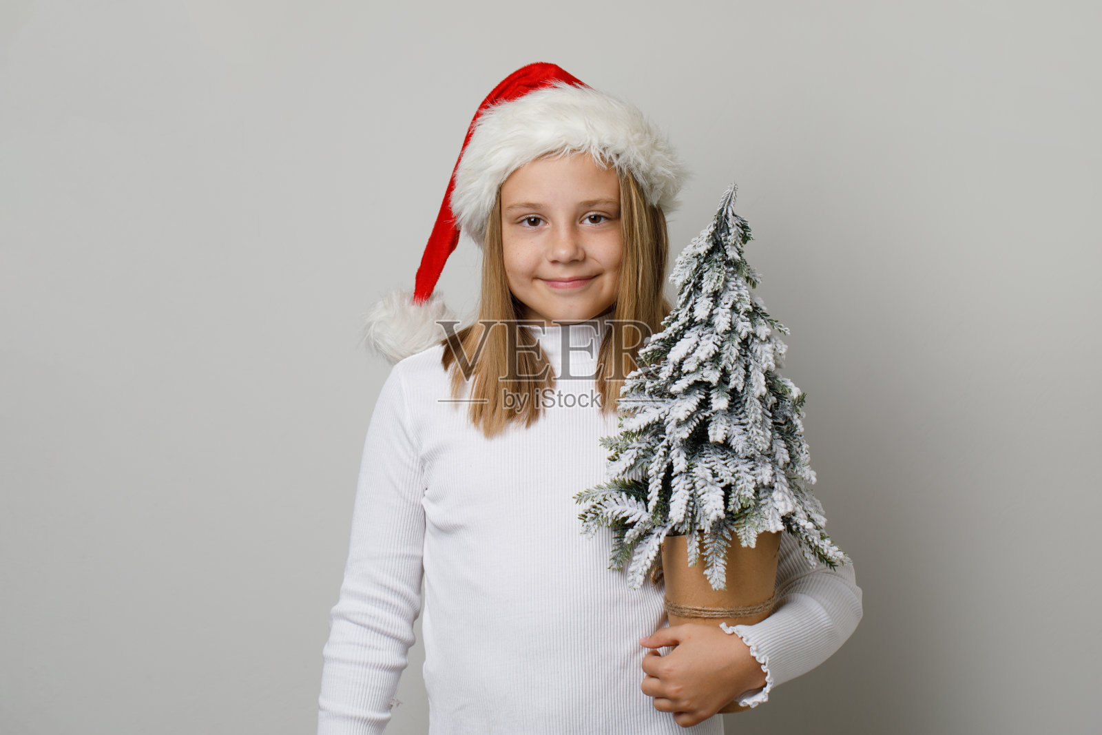 戴着圣诞老人帽，穿着白色t恤的小女孩手持圣诞树。圣诞小孩10岁的肖像照片摄影图片