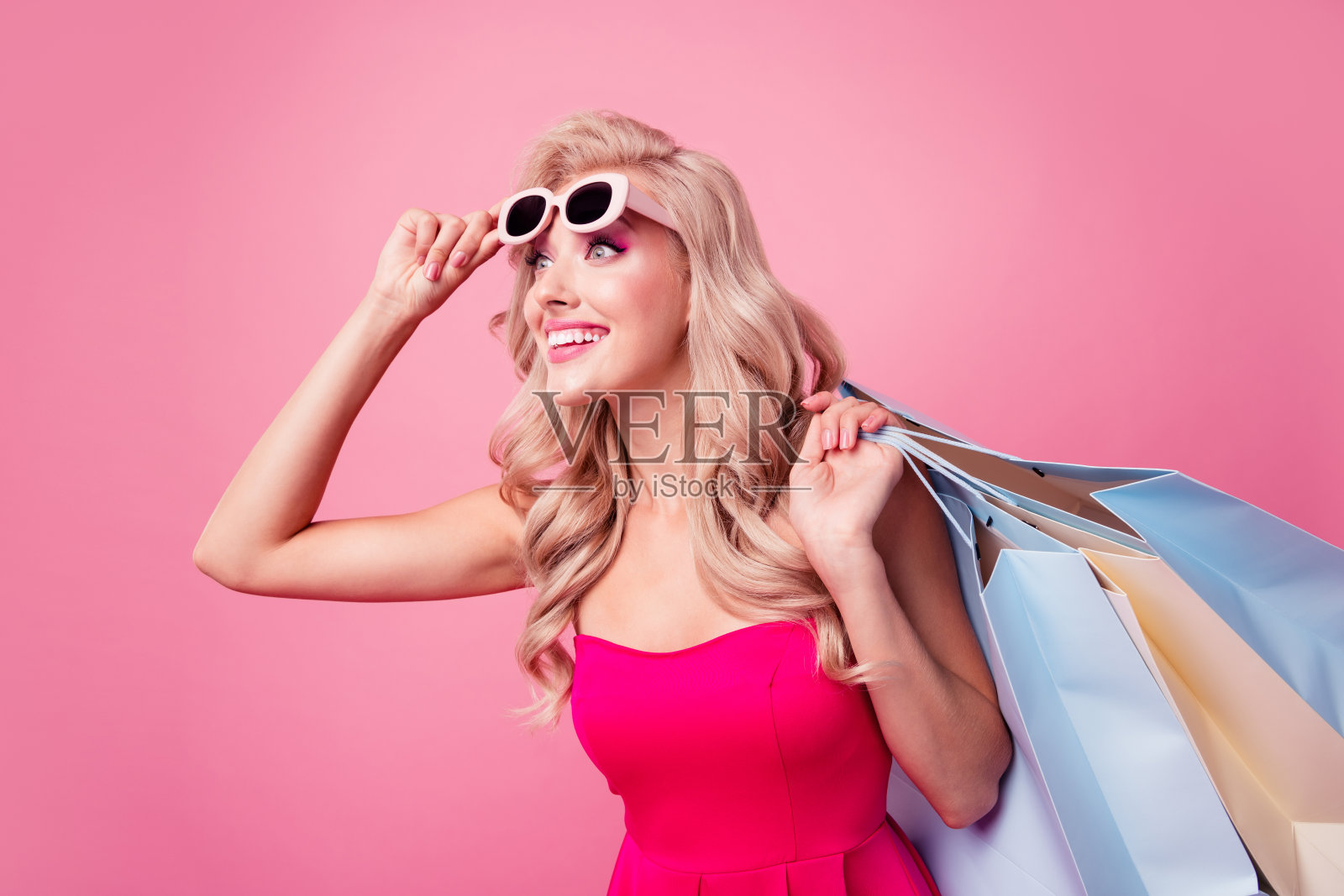 照片中，令人震惊的女孩拿着购物袋，触摸太阳镜，在粉红色的背景下，看起来空无一物照片摄影图片