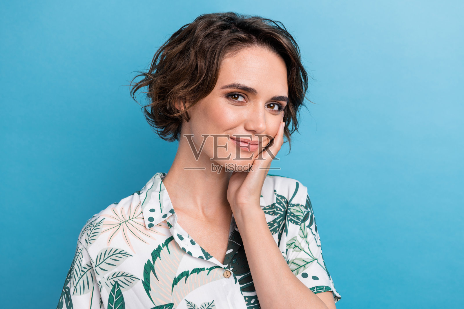 漂亮可爱的开朗女孩的照片，鲍勃发型，穿着彩色衬衫，手掌放在颧骨上，孤立在蓝色背景上照片摄影图片