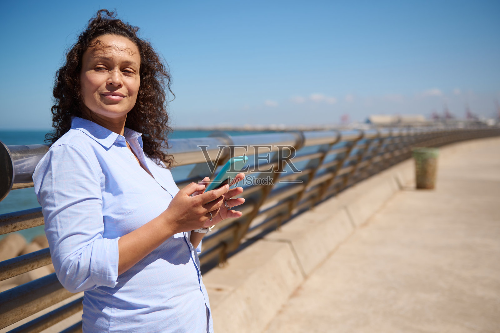 穿着便服，手持智能手机，微笑着望向一边，站在海景的背景下。照片摄影图片