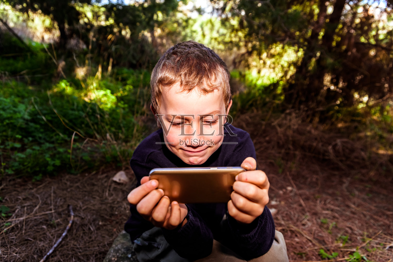 在森林里用智能手机看视频的男孩，表情非常开心。照片摄影图片