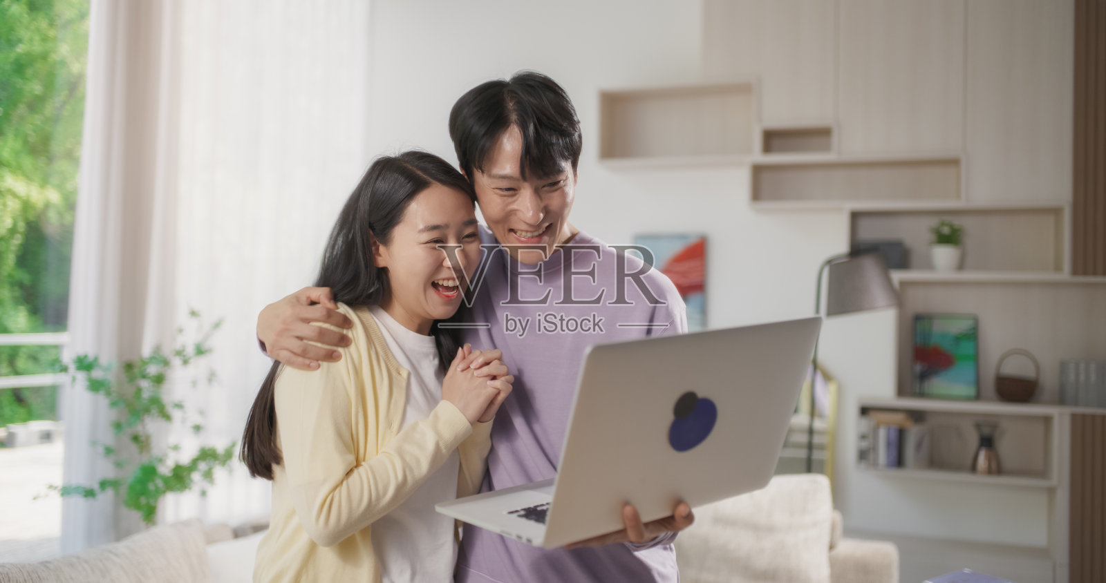 年轻可爱的韩国夫妇站在时尚的客厅里，在家里使用笔记本电脑。男人和女人等待成功的结果，快乐，欢呼和庆祝胜利照片摄影图片