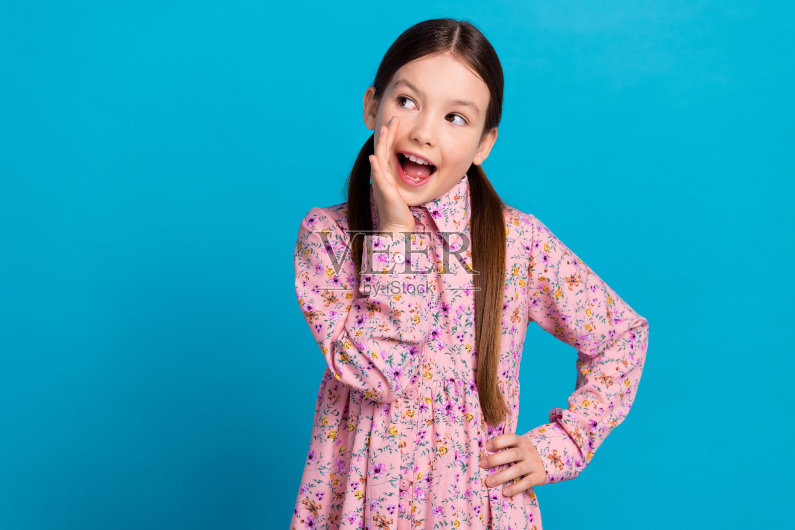 可爱的小女孩的照片肖像分享秘密的手盖嘴穿时髦的粉红色印花衣服孤立的蓝色背景照片摄影图片