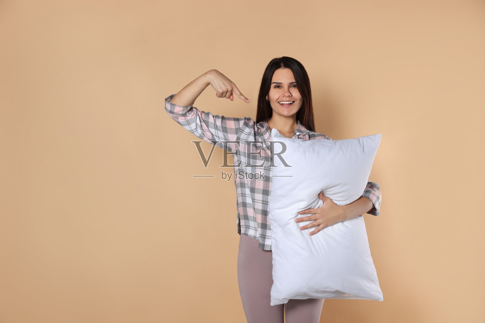 快乐的年轻女子指着米色背景上柔软的枕头，为文字留出空间照片摄影图片