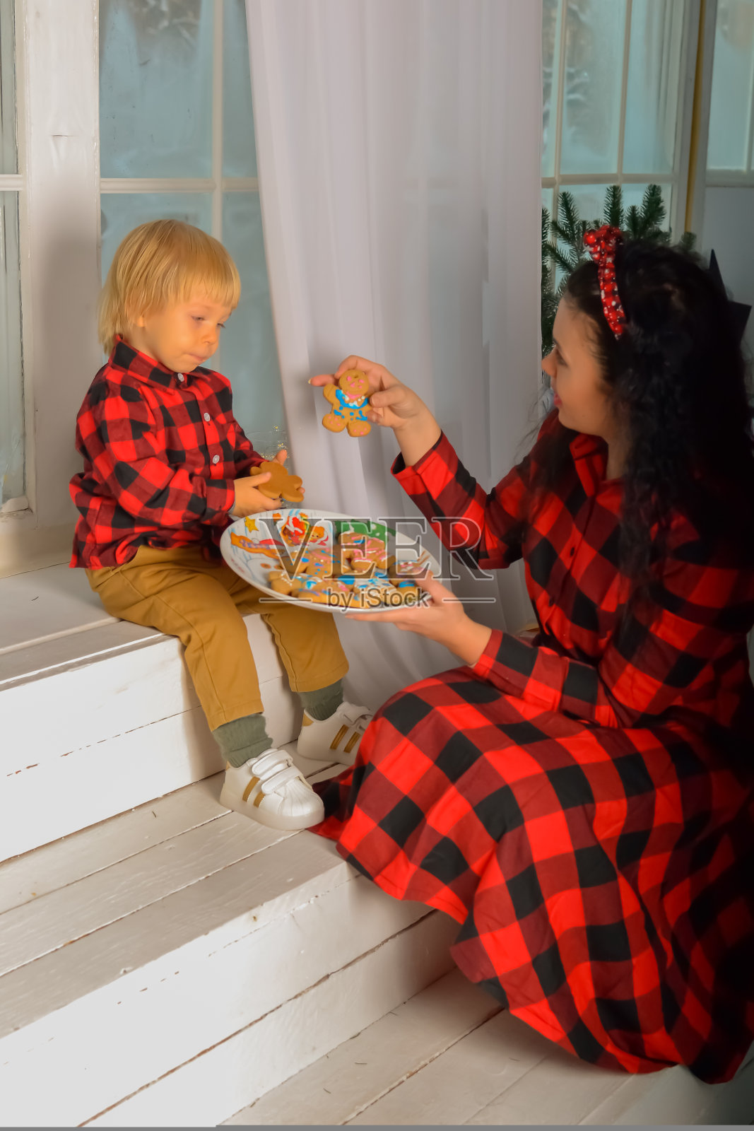 妈妈和儿子在窗边吃圣诞饼干。母亲和儿子穿着一模一样的圣诞服装，坐在窗台上，手里拿着一块圣诞饼干。家人看。圣诞奇迹。家庭圣诞节概念照片摄影图片