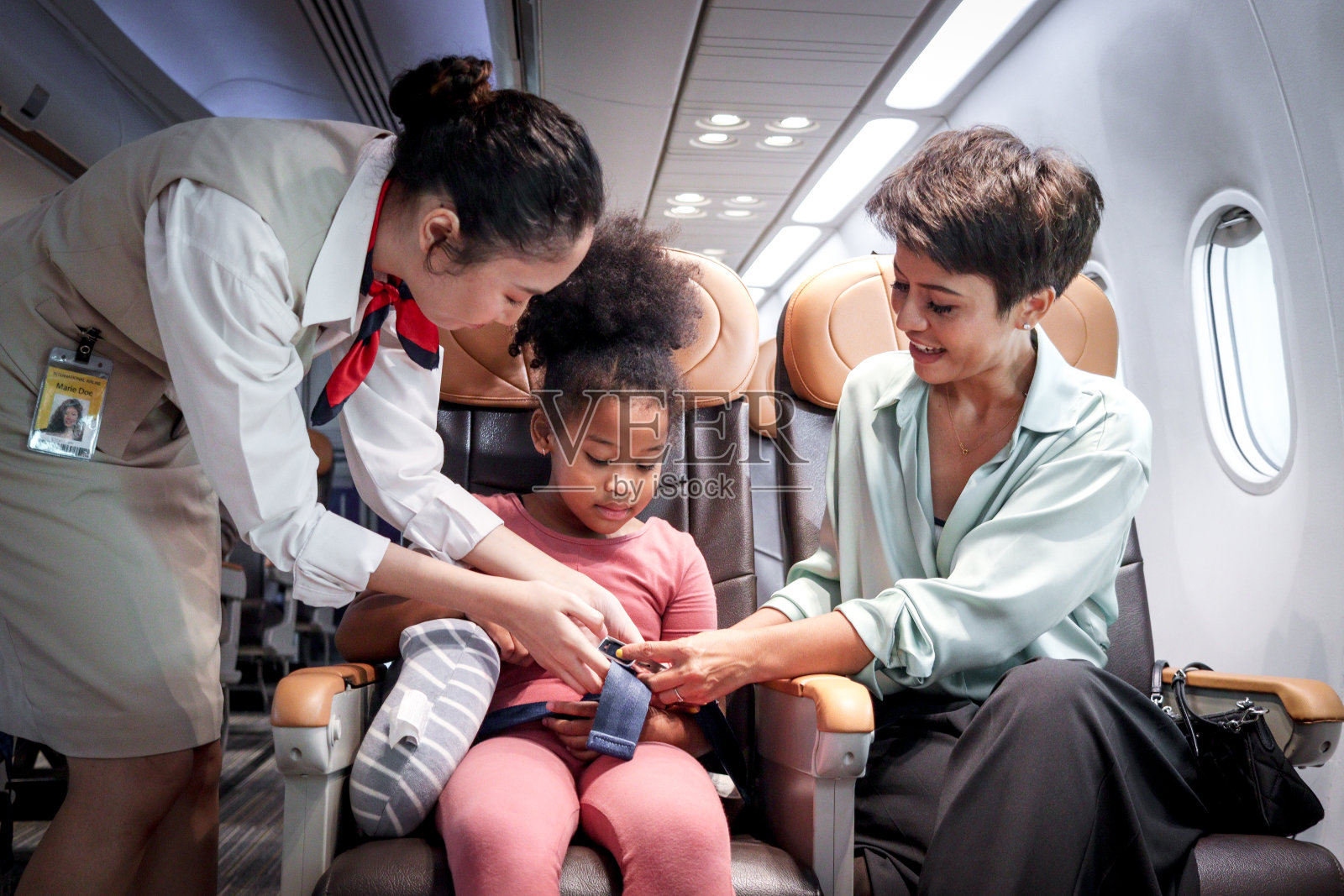 友好的空中小姐在飞机上照顾小孩乘客，空乘人员在飞机座位上就教卷发非洲女孩使用安全带，小孩乘飞机旅行，航空运输照片摄影图片