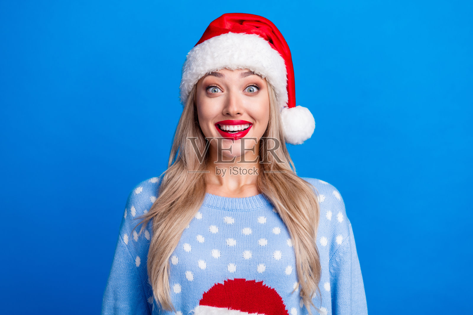 漂亮的金发女孩的照片肖像激动印象深刻的牙齿微笑穿着毛衣圣诞帽孤立的蓝色背景照片摄影图片