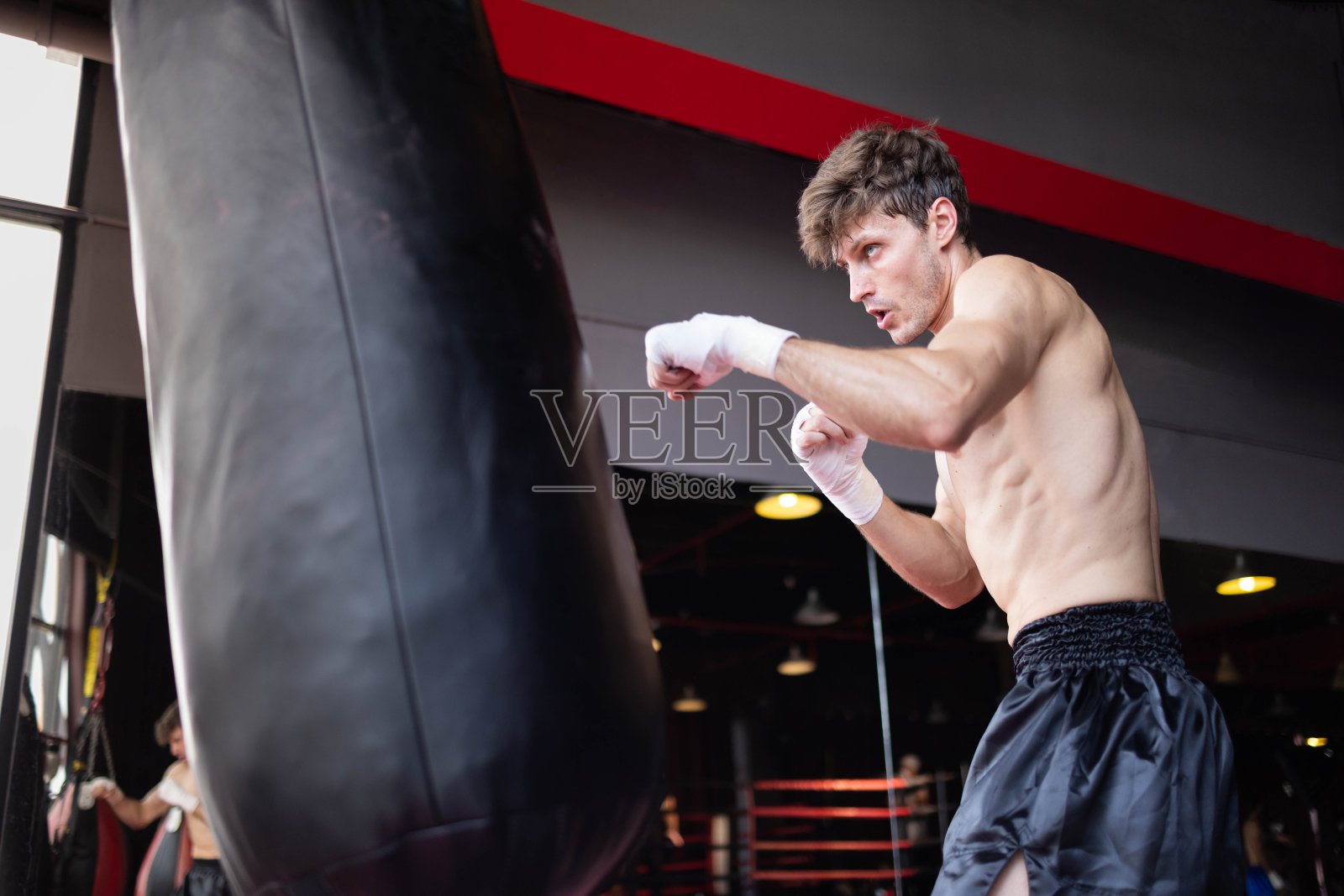图为，身穿运动服，戴着拳击手套的活跃的白人职业运动员在健身房或健身俱乐部用拳击沙袋或拳击沙袋进行训练。照片摄影图片