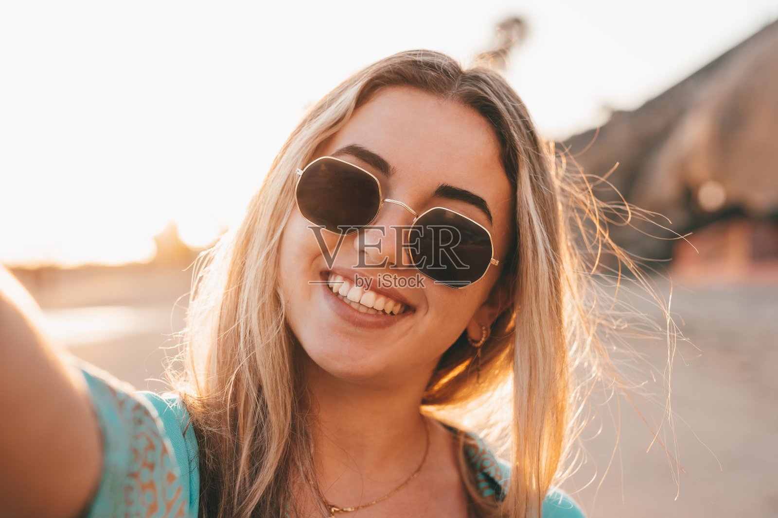 一个年轻美丽迷人的金发女郎手持相机在沙滩上自拍，享受户外自由时光，背景是日落。照片摄影图片