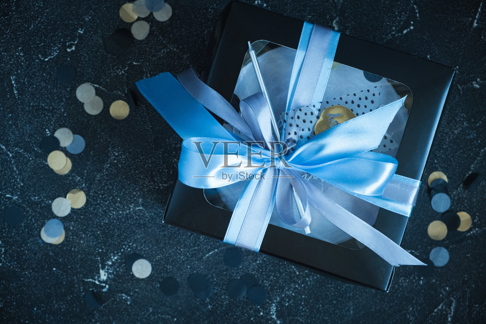 黑色礼盒里的小便当蛋糕，背景深蓝色。平的。生日礼物装在一个黑盒子里，用蓝丝带系着蝴蝶结照片摄影图片