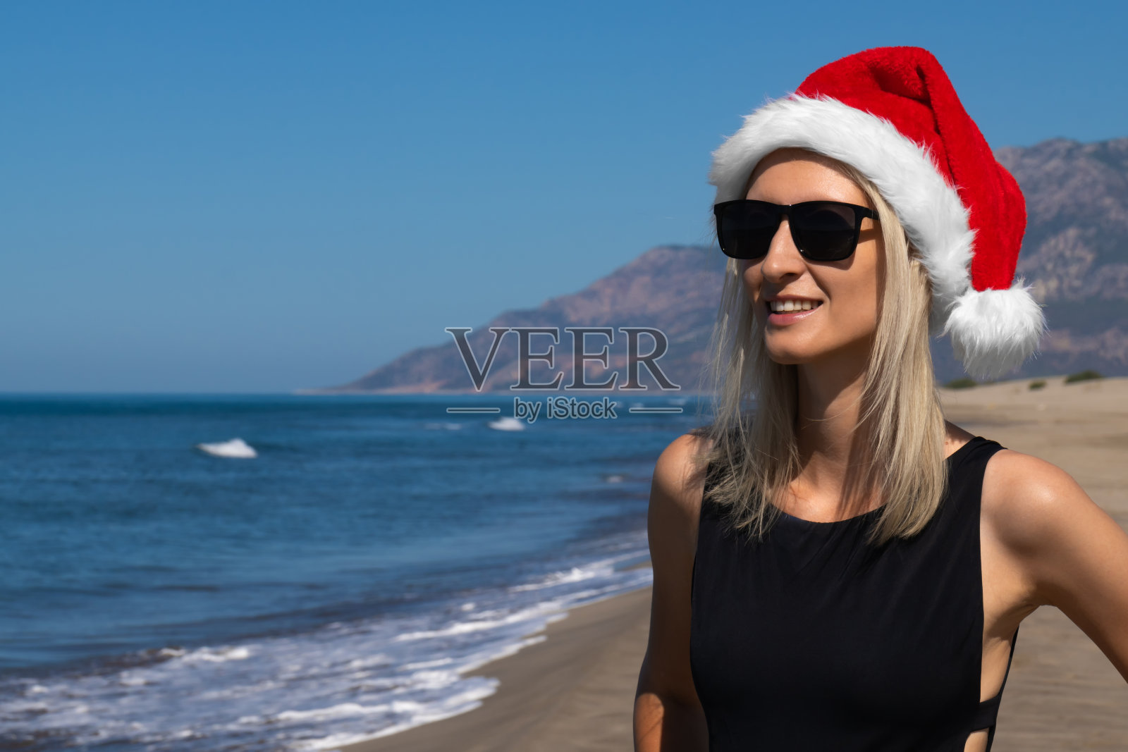 年轻的金发女子穿着黑色泳衣，戴着圣诞老人帽，戴着太阳镜，在阳光明媚的日子里快乐而美丽地站在海边和山上。圣诞假期照片摄影图片