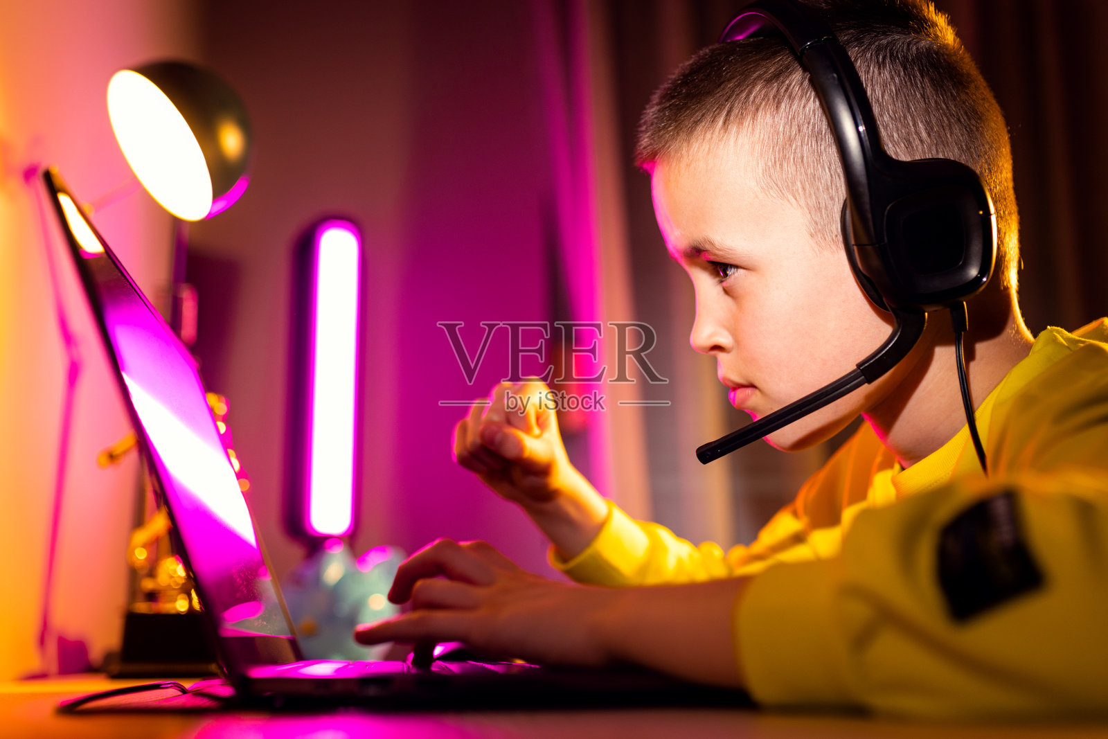 年轻兴奋的男孩玩家戴着耳机在家里的笔记本电脑上玩视频游戏。照片摄影图片