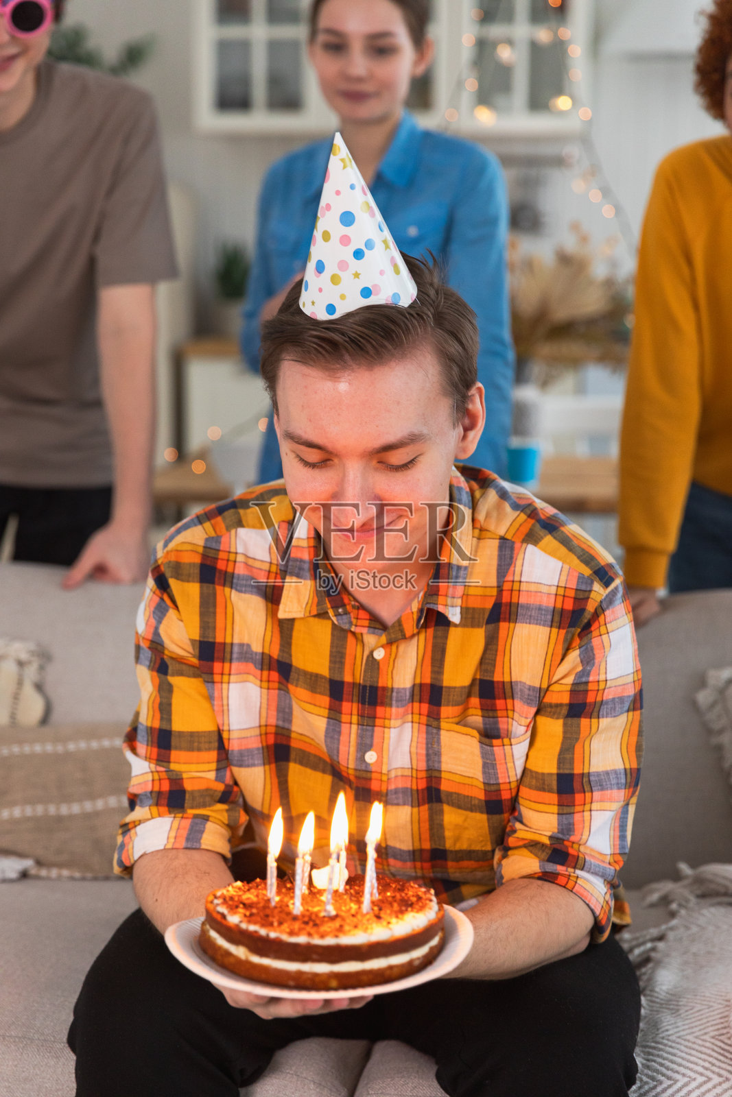 许个愿。戴着派对帽的男子吹灭了生日蛋糕上燃烧的蜡烛。生日派对快乐。一群朋友祝他生日快乐。人们在家里开派对庆祝生日。照片摄影图片