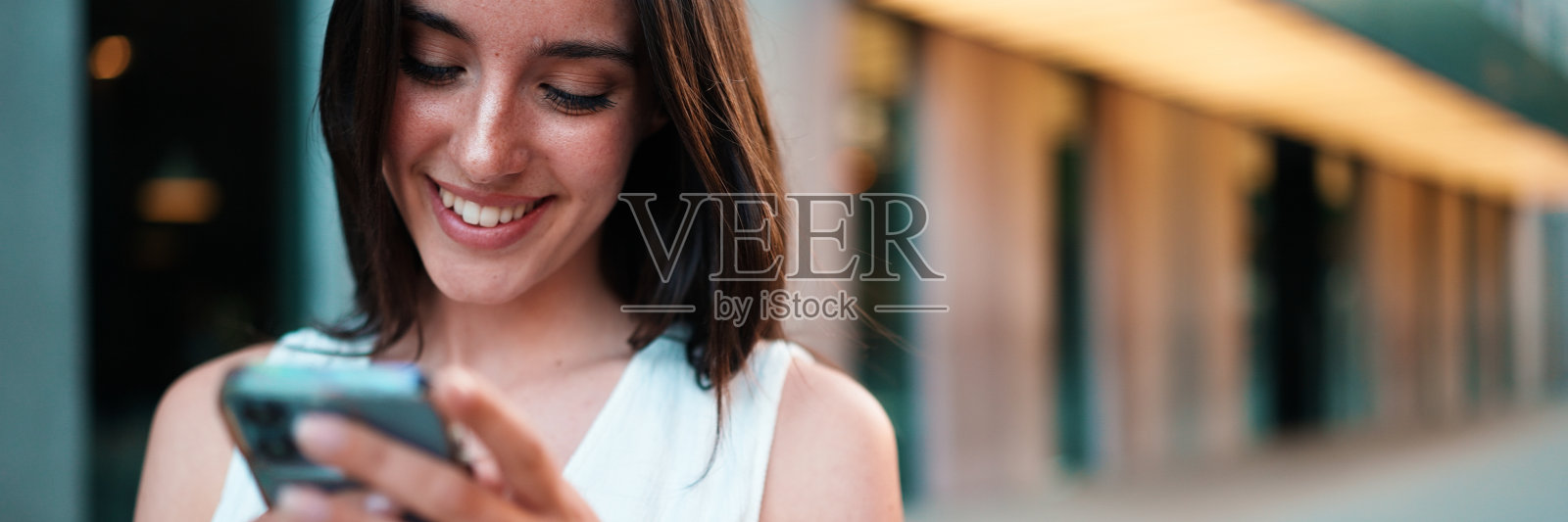 一个长着雀斑、深色蓬松头发、穿着白色上衣的美女，手里拿着智能手机走在街上。女孩在现代城市背景下使用手机照片摄影图片