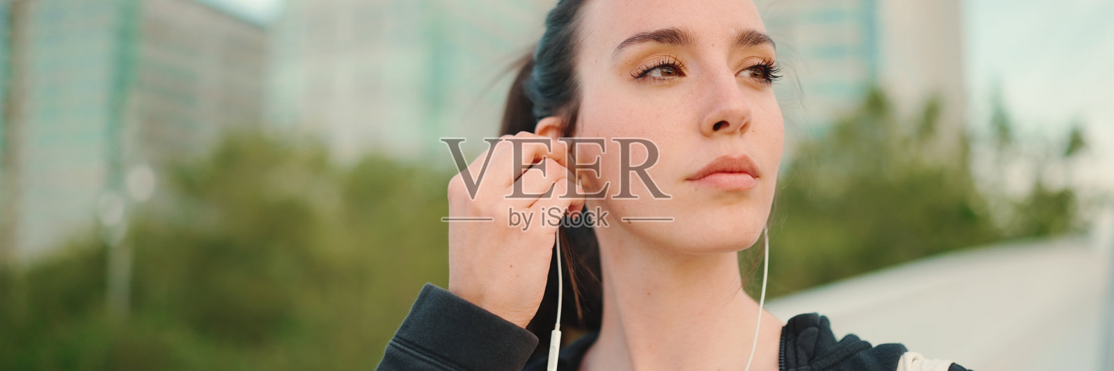 一个长着雀斑、扎着长马尾、穿着黑色运动连帽衫、戴着有线耳机的年轻女子一边听着音乐，一边沿着桥走着照片摄影图片