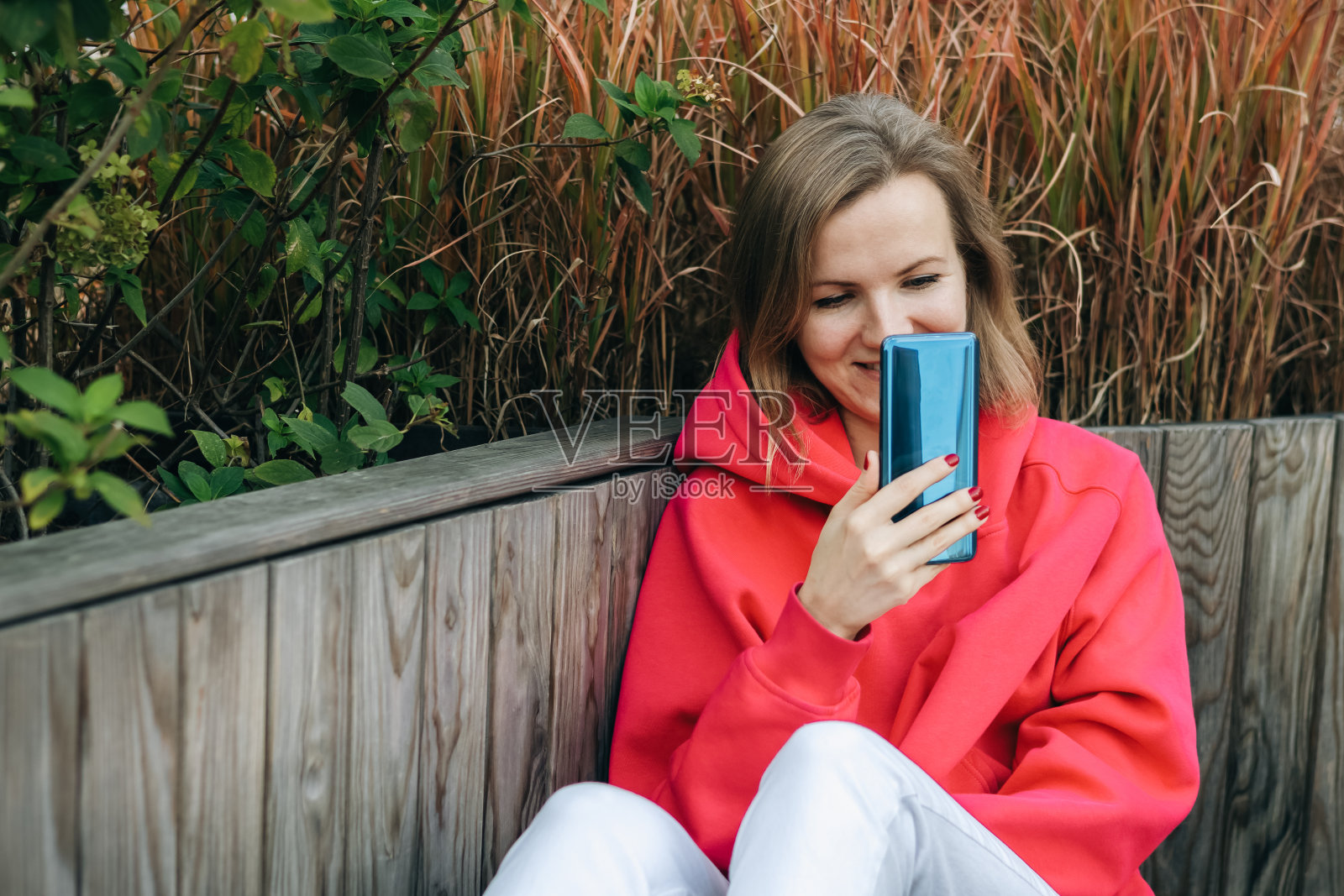 穿着红色连帽衫和白色运动鞋的美女正在户外长凳上用智能手机打视频电话。照片摄影图片