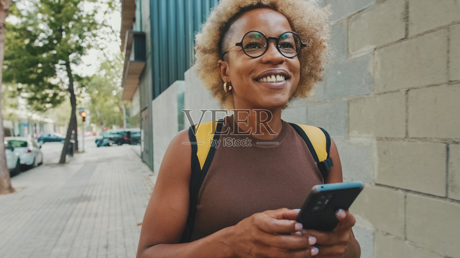 微笑的年轻女子戴着眼镜，穿着棕色上衣，肩上背着背包，走在街上，拿着智能手机，发短信，在社交媒体上和朋友聊天照片摄影图片