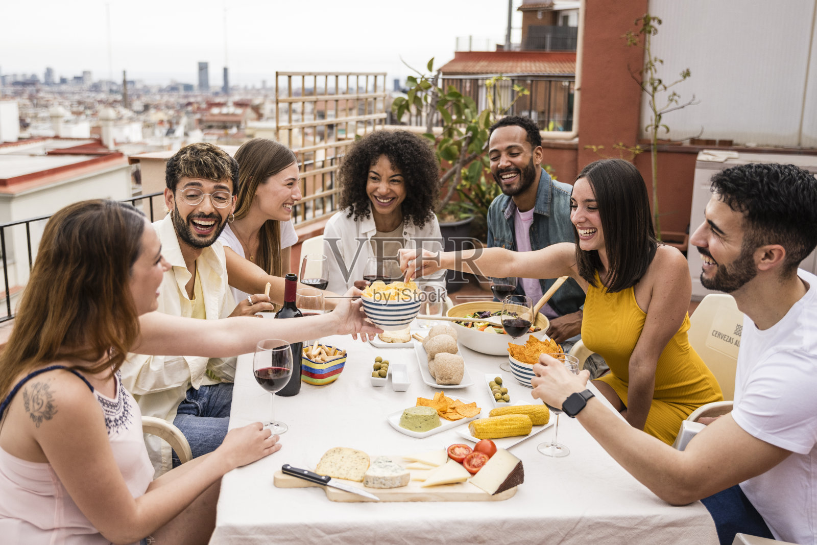 一群不同的朋友在露台上吃饭喝酒。年轻而放松的人们在屋顶晚宴上玩得很开心。照片摄影图片