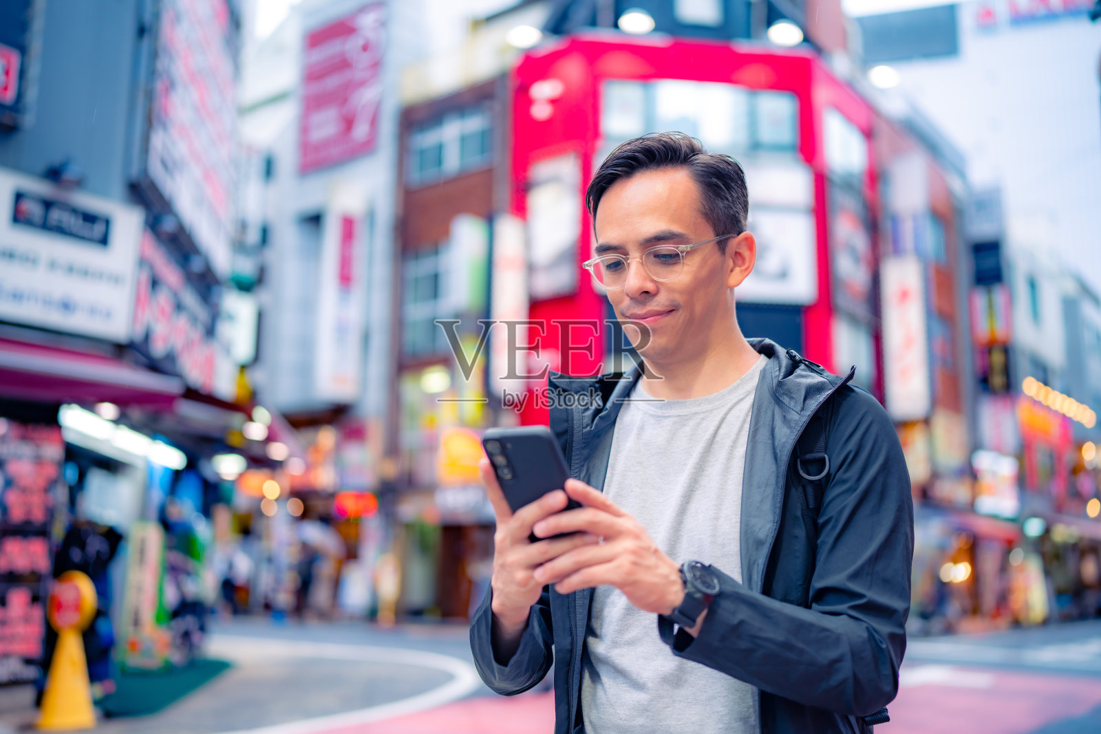 穿着休闲服，戴着眼镜，快乐的西班牙裔年轻男性走在日本东京的新宿附近，在白天浏览智能手机照片摄影图片