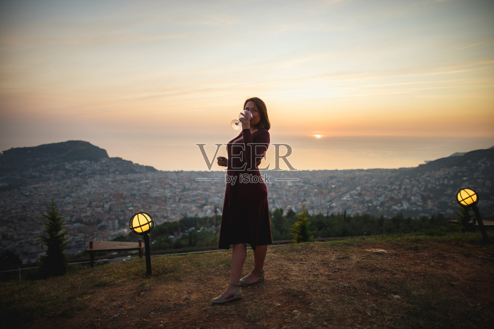 夕阳下的山间，一名身穿酒红色长裙的性感成熟女子从杯中啜饮红酒照片摄影图片