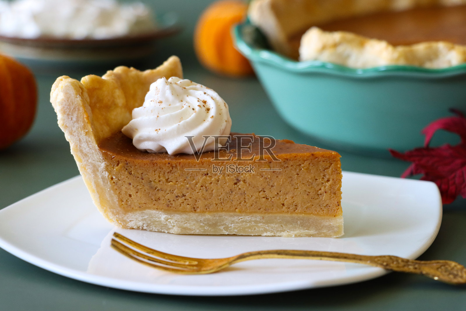 特写图片:一片新鲜出炉的南瓜派，上面有搅打过的奶油，馅饼锡盒里装着自由形状的有凹槽的派皮，里面装满了南瓜泥，金色的甜点叉子，绿色背景，重点放在前景上照片摄影图片