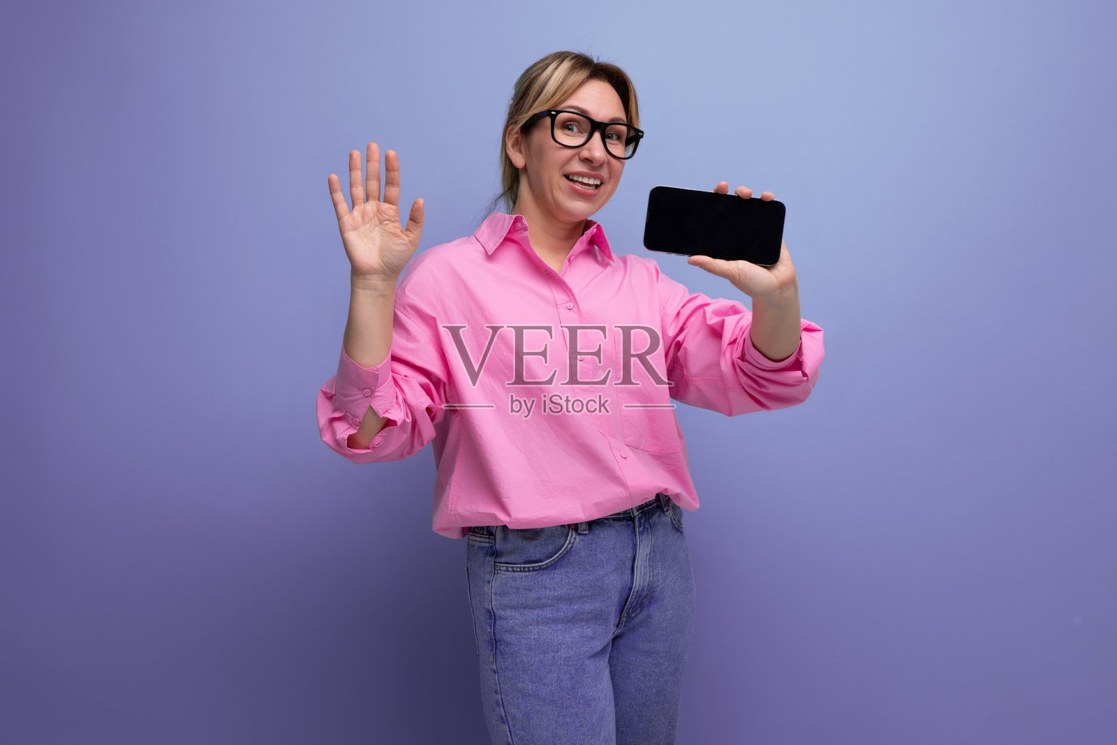 年轻成功的白种金发白领女性穿着粉红色的衬衫和牛仔裤，戴着眼镜，梳着马尾辫，在电话里播放广告照片摄影图片